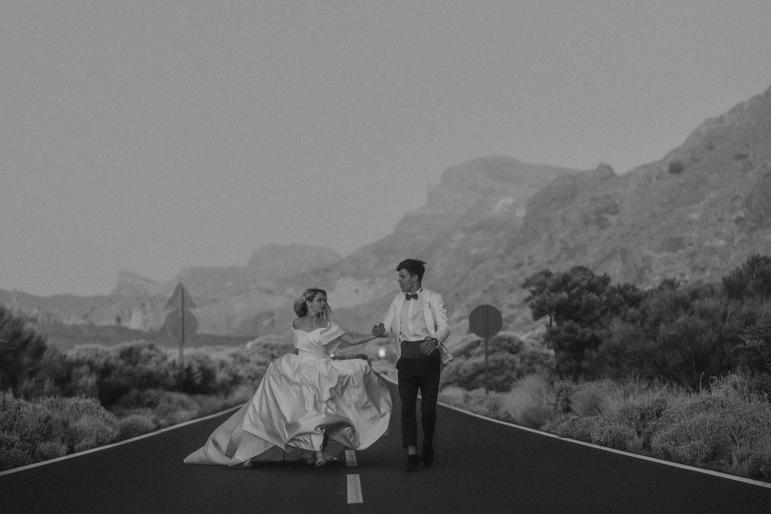 Black and white photo of a bride and groom walking hand in hand on an empty road with mountains in the background.