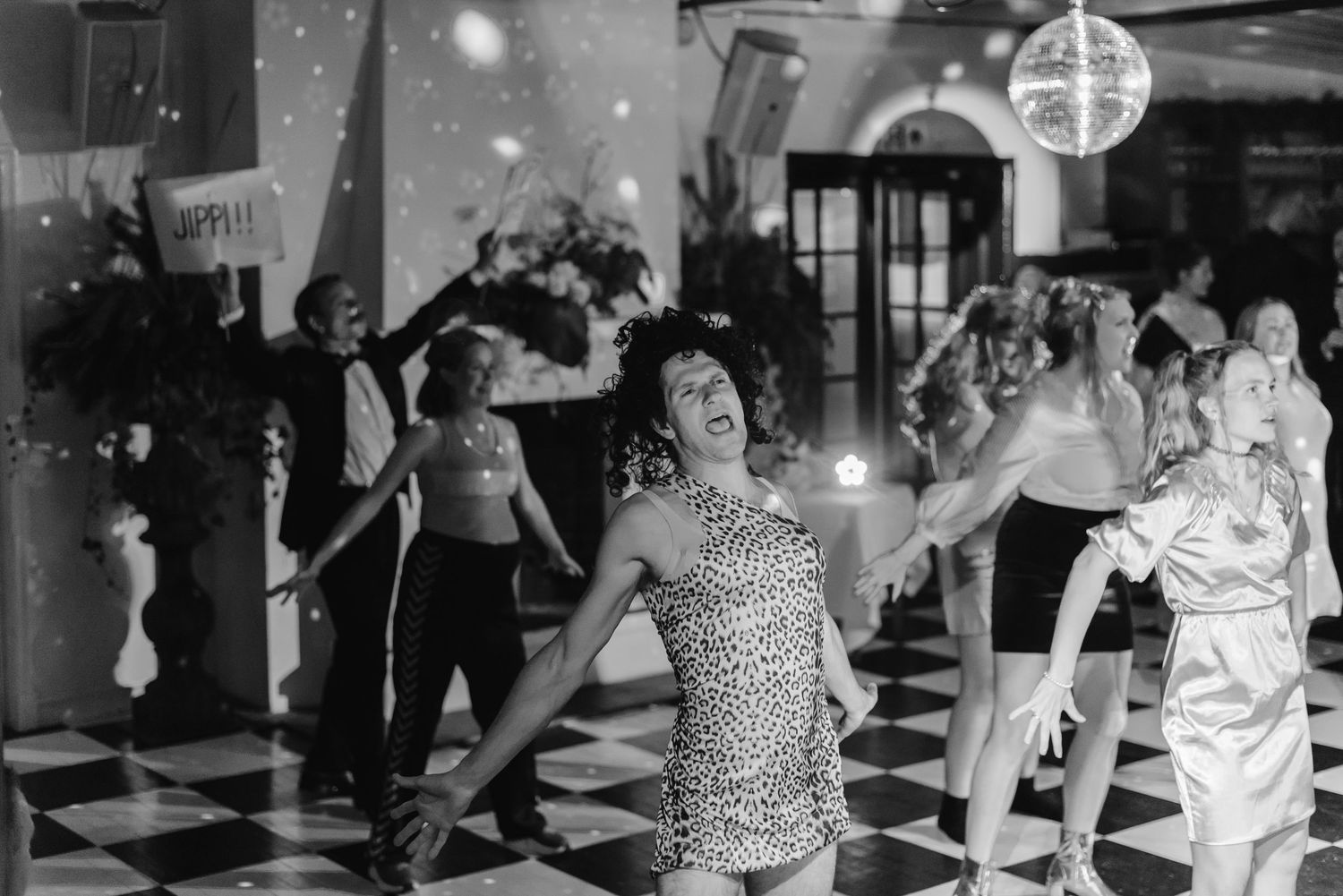 People dancing at a retro disco party with a mirror ball, checkered floor, and festive decorations in black and white photo.