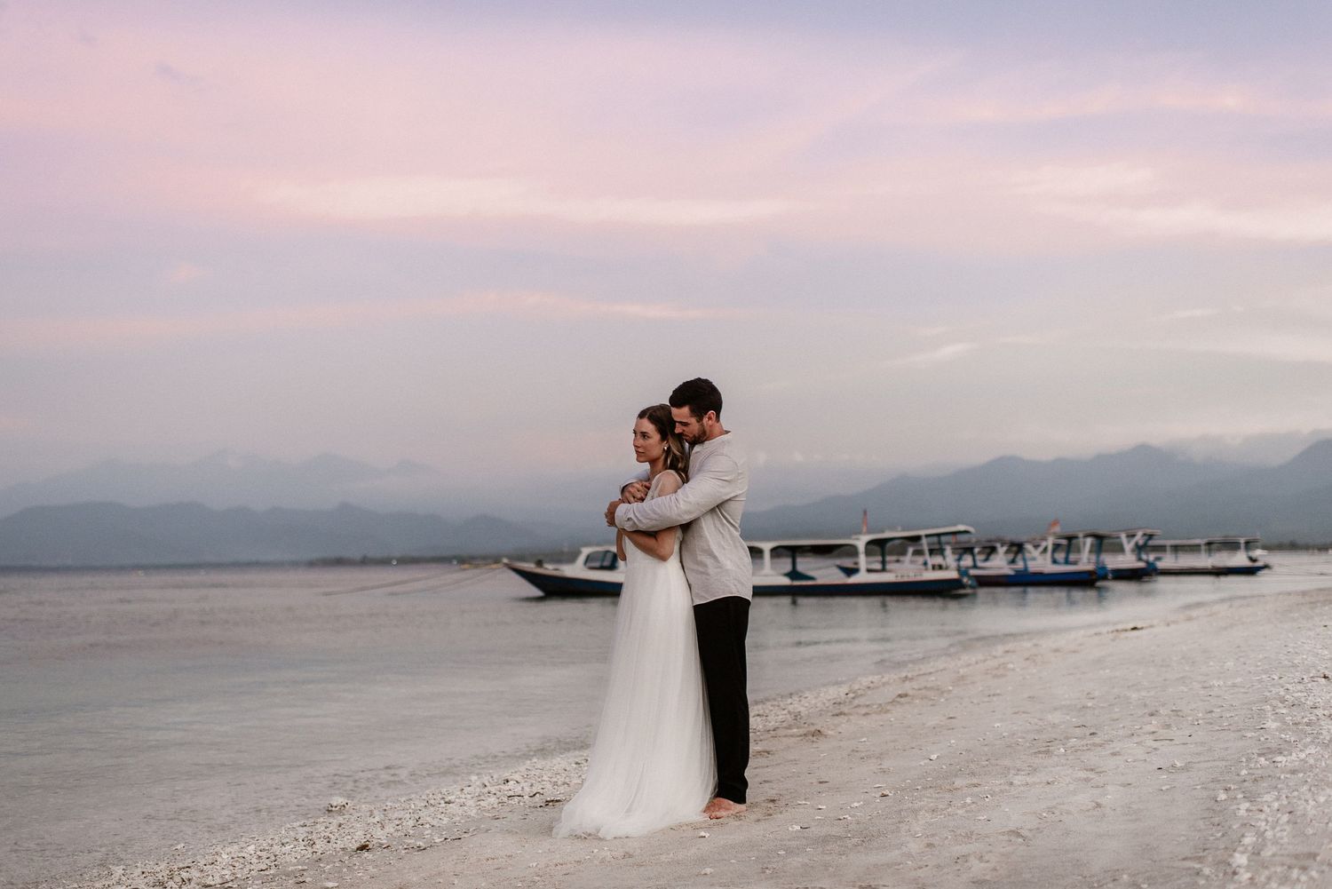 Couple embracing on a white sand beach at sunset with a boat and mountains in the background.