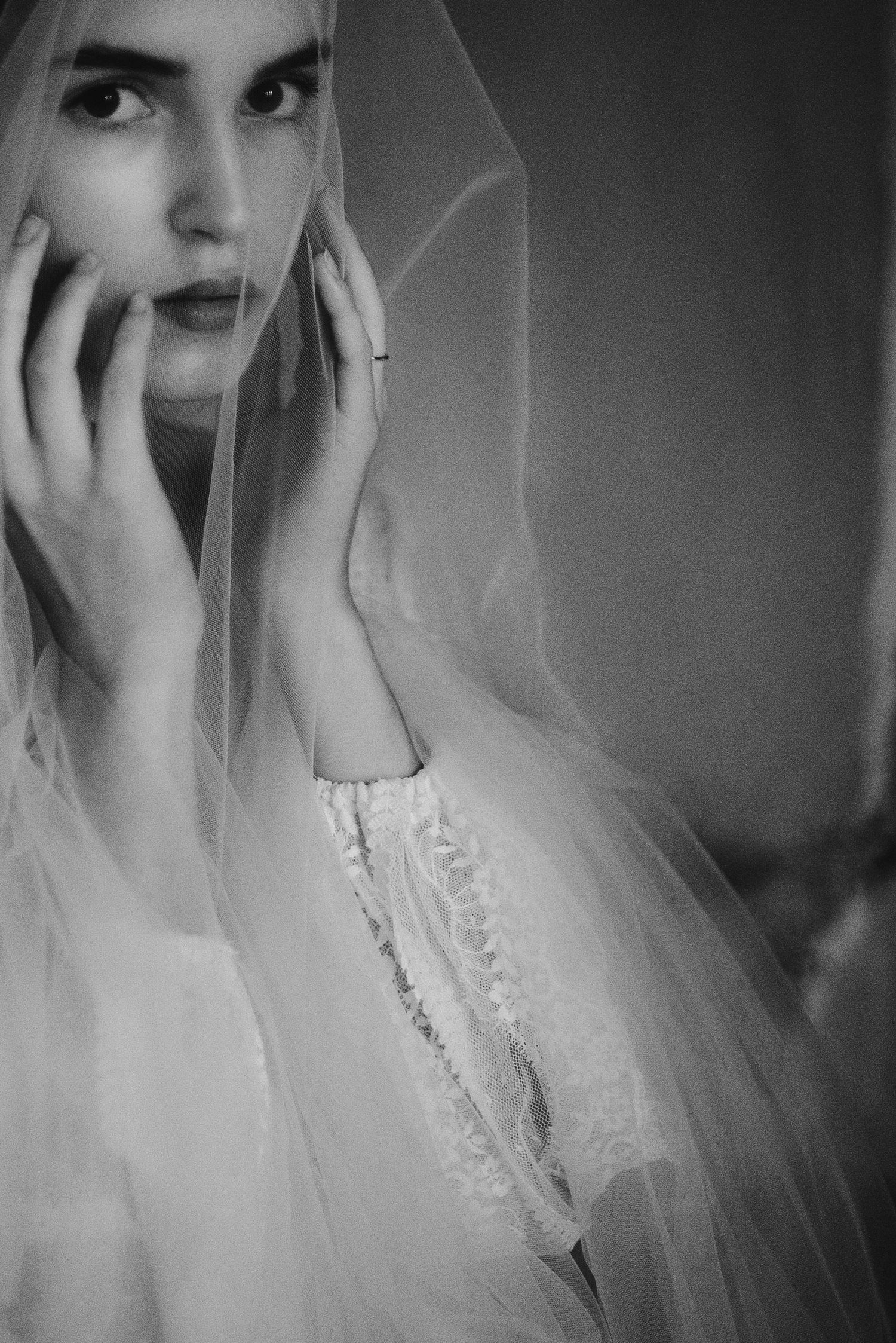 Black and white portrait of a bride in a lace wedding dress and veil with hands near face in dramatic lighting.