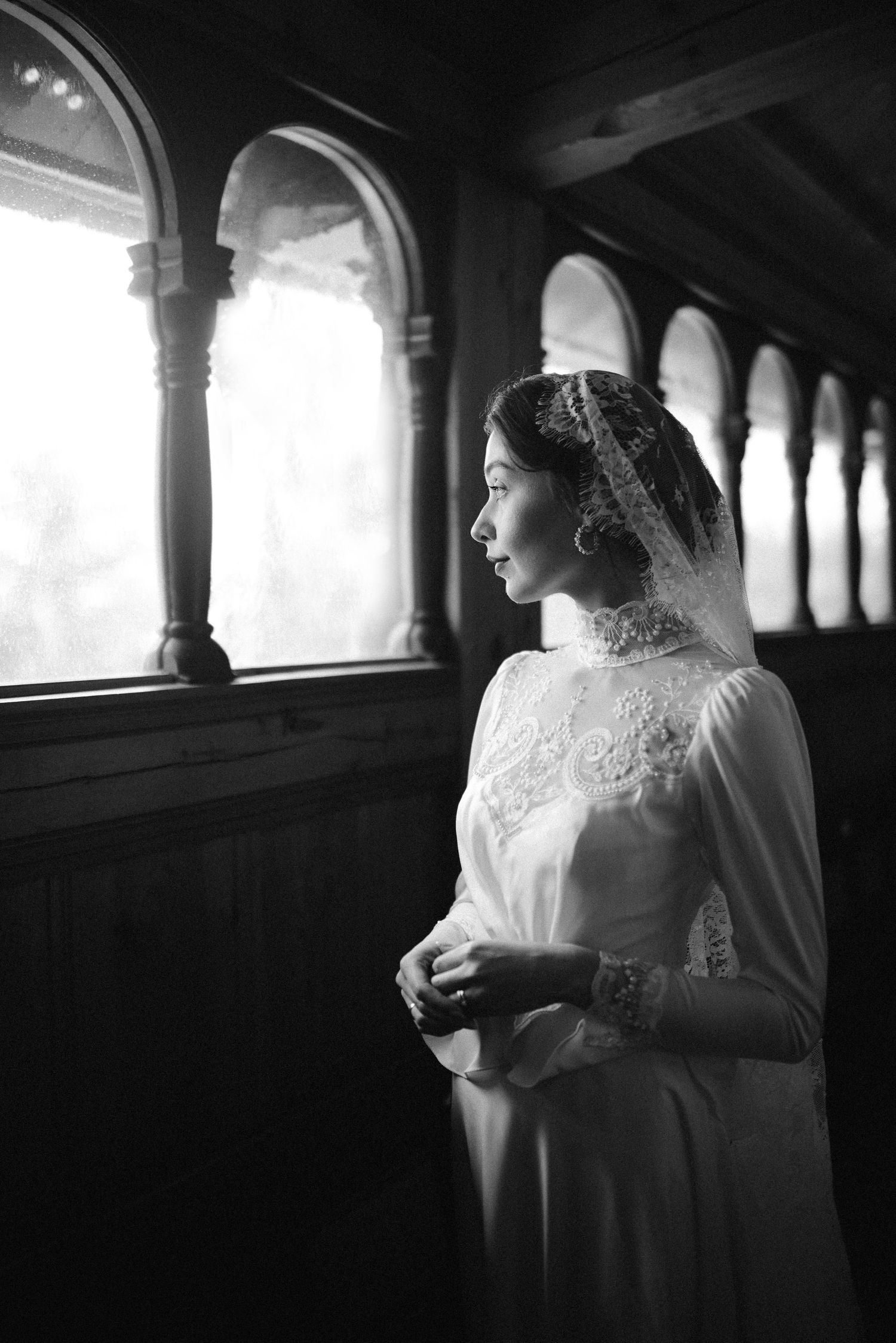 Black and white photo of a bride in a lace veil and white gown standing by arched windows in a historic building.