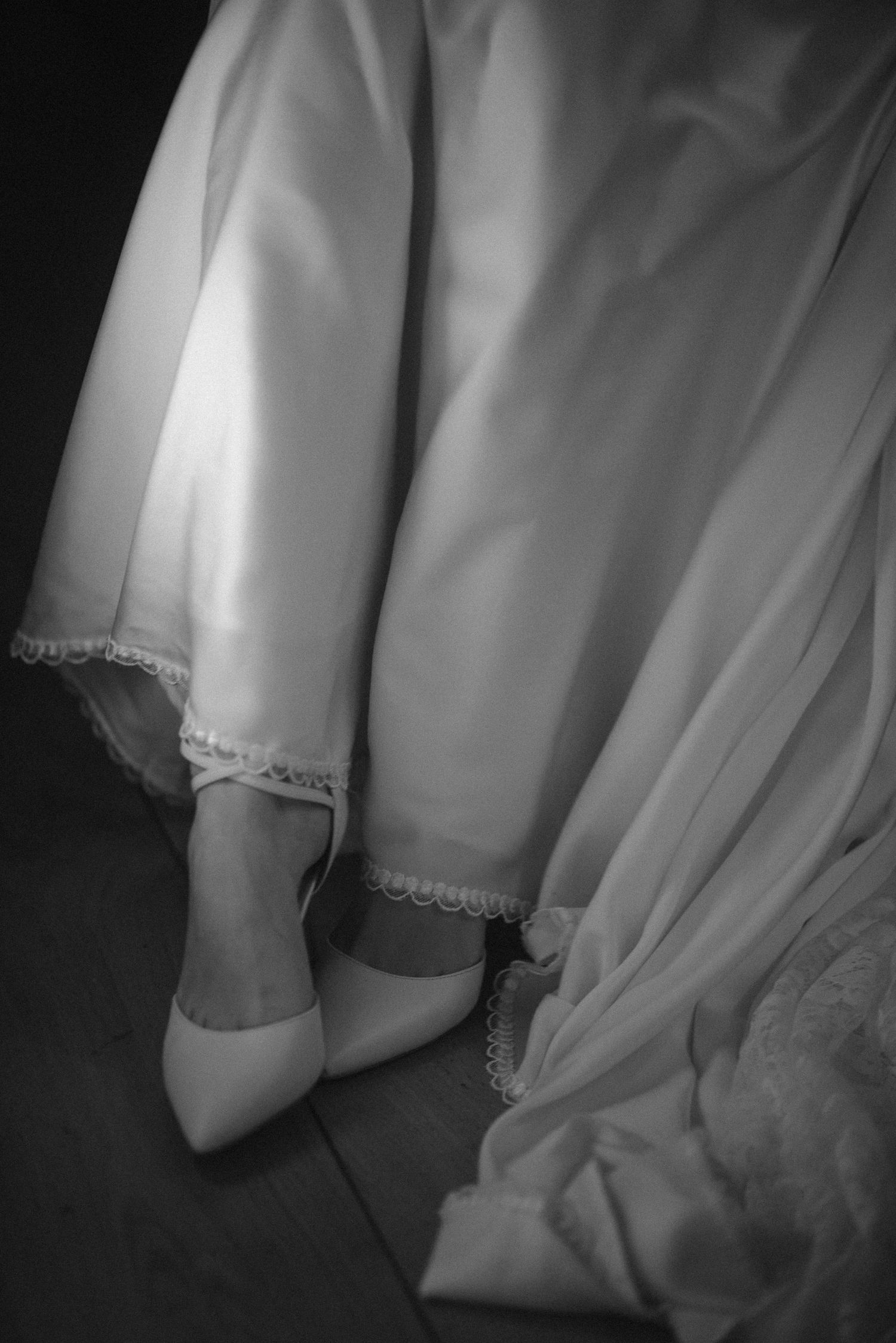 Black and white photo of a bride in a white wedding gown wearing elegant heels on a wooden floor.