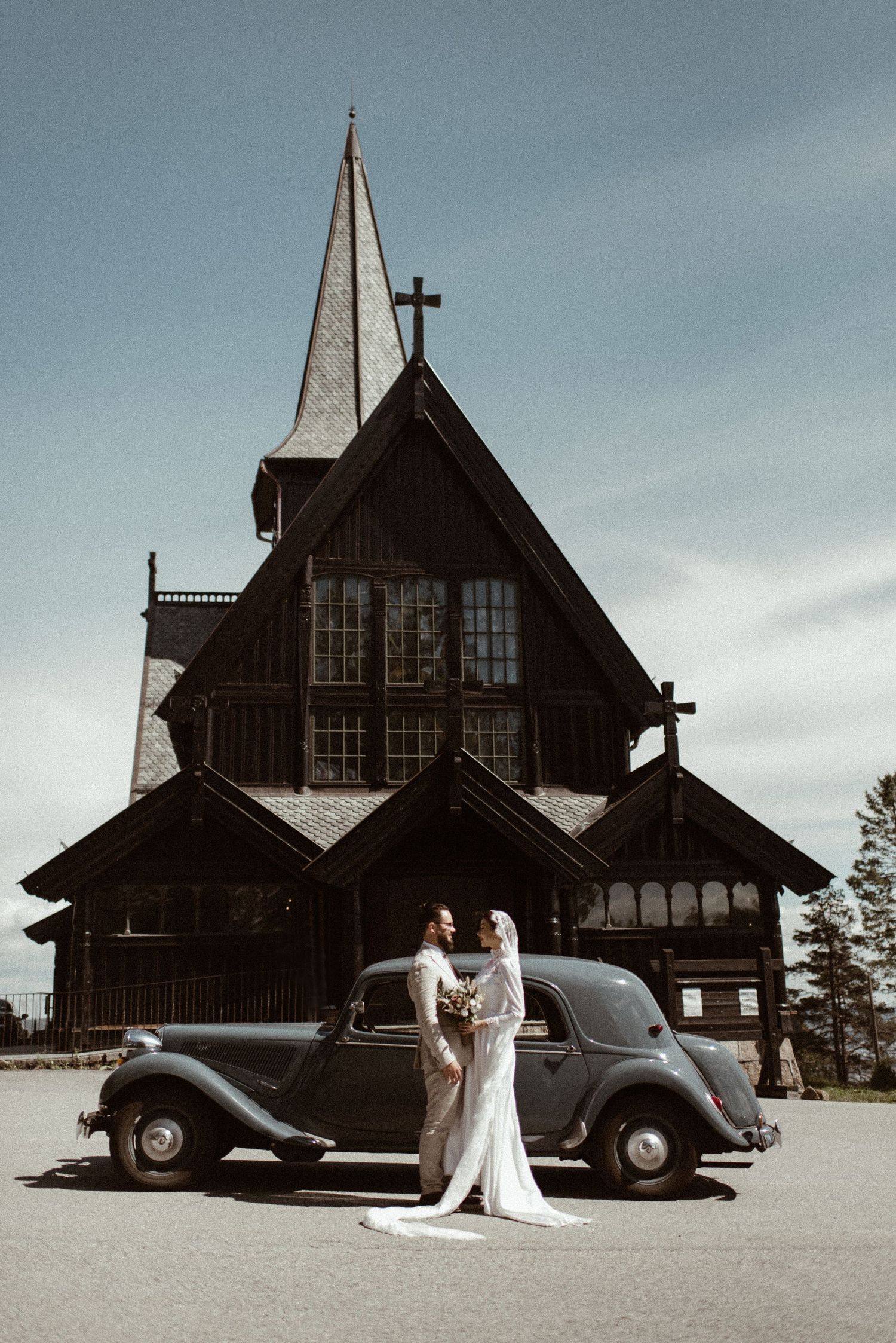 Bride and groom pose beside a vintage car in front of a dark wooden stave church under a clear sky.