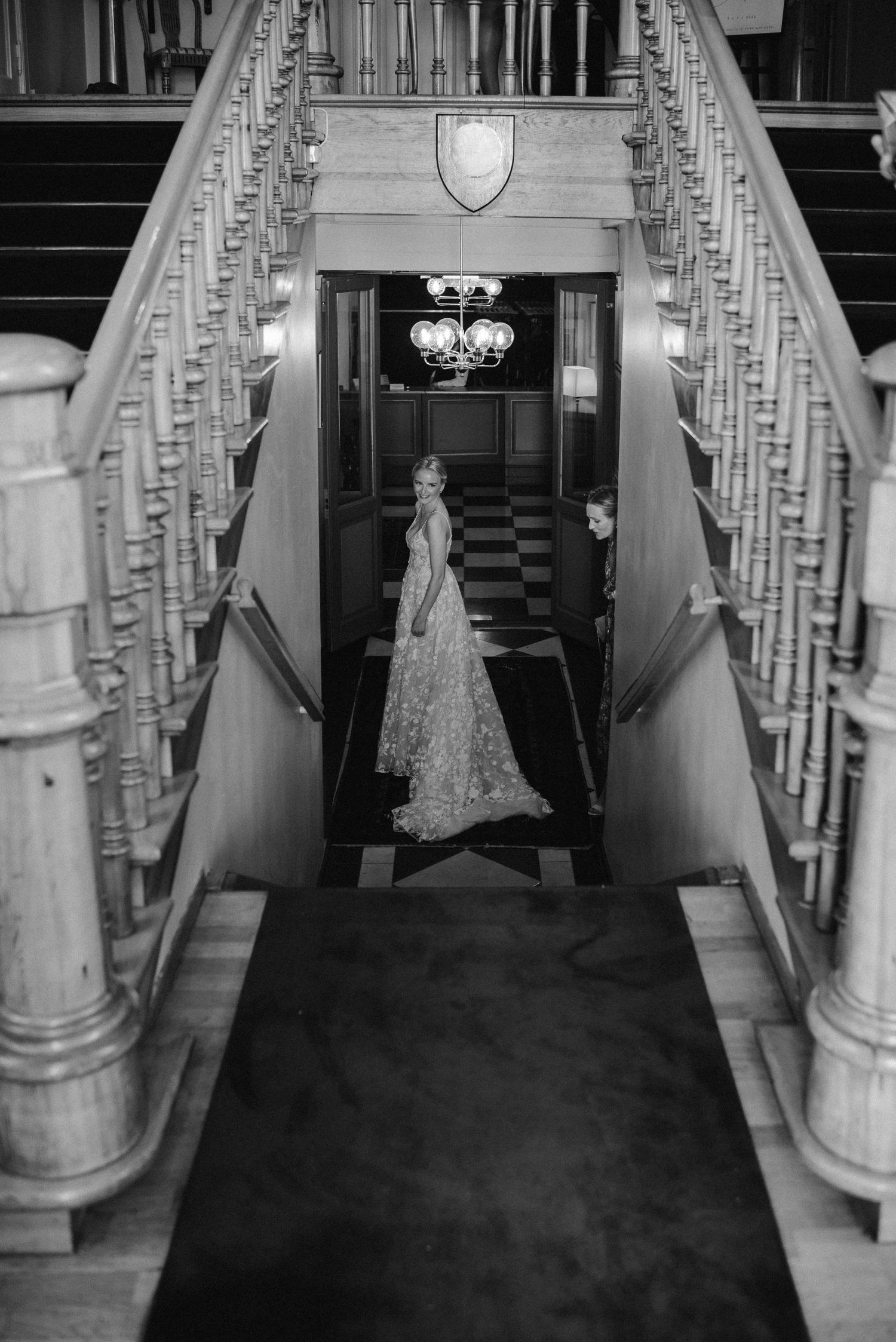 Black and white photo of a bride in a lace gown standing at the base of an elegant grand staircase.