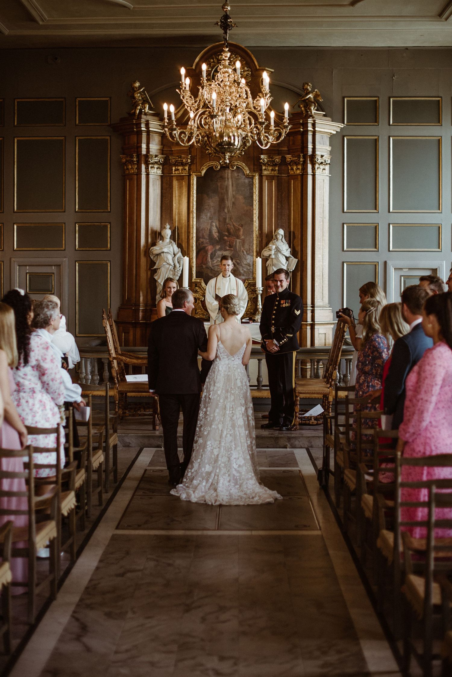 Elegant wedding ceremony in ornate chapel with chandelier, bride in lace gown, groom in military uniform, seated guests.
