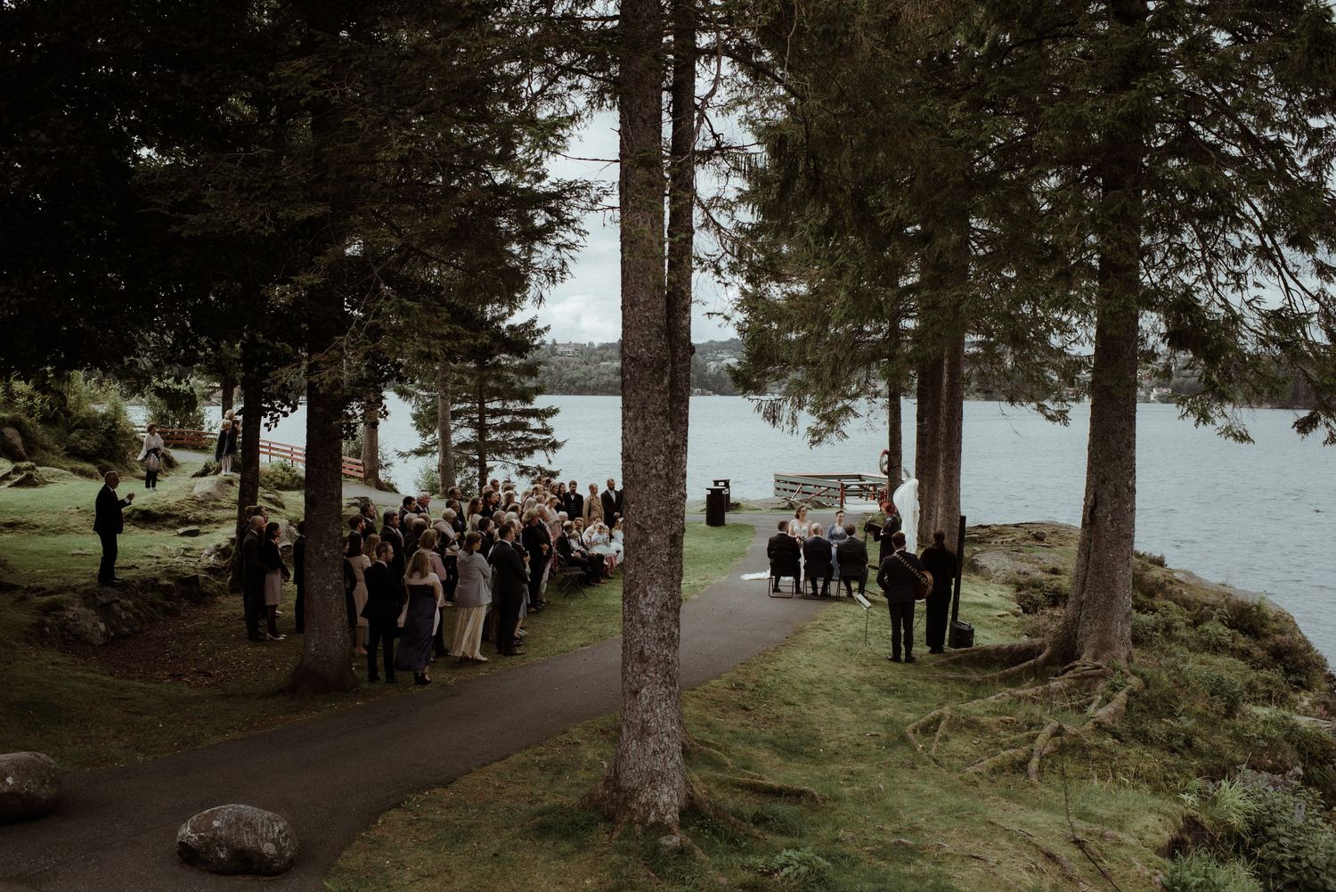 Outdoor wedding ceremony among tall pine trees beside a serene lake with guests gathered on a rocky path.