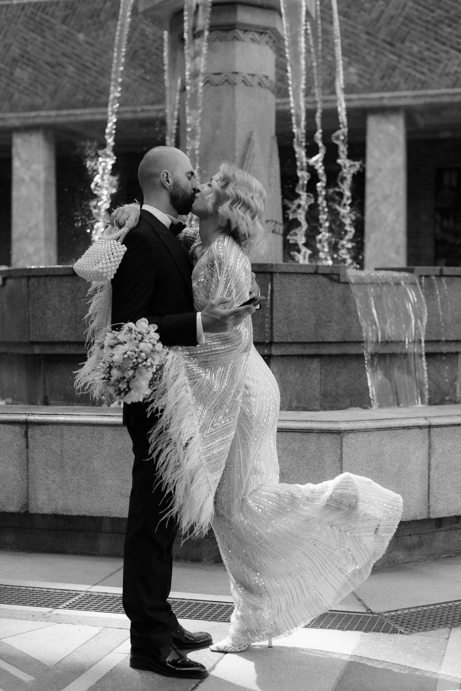 Black and white photo of a bride and groom kissing by a fountain, bride in feathered gown holding bouquet.