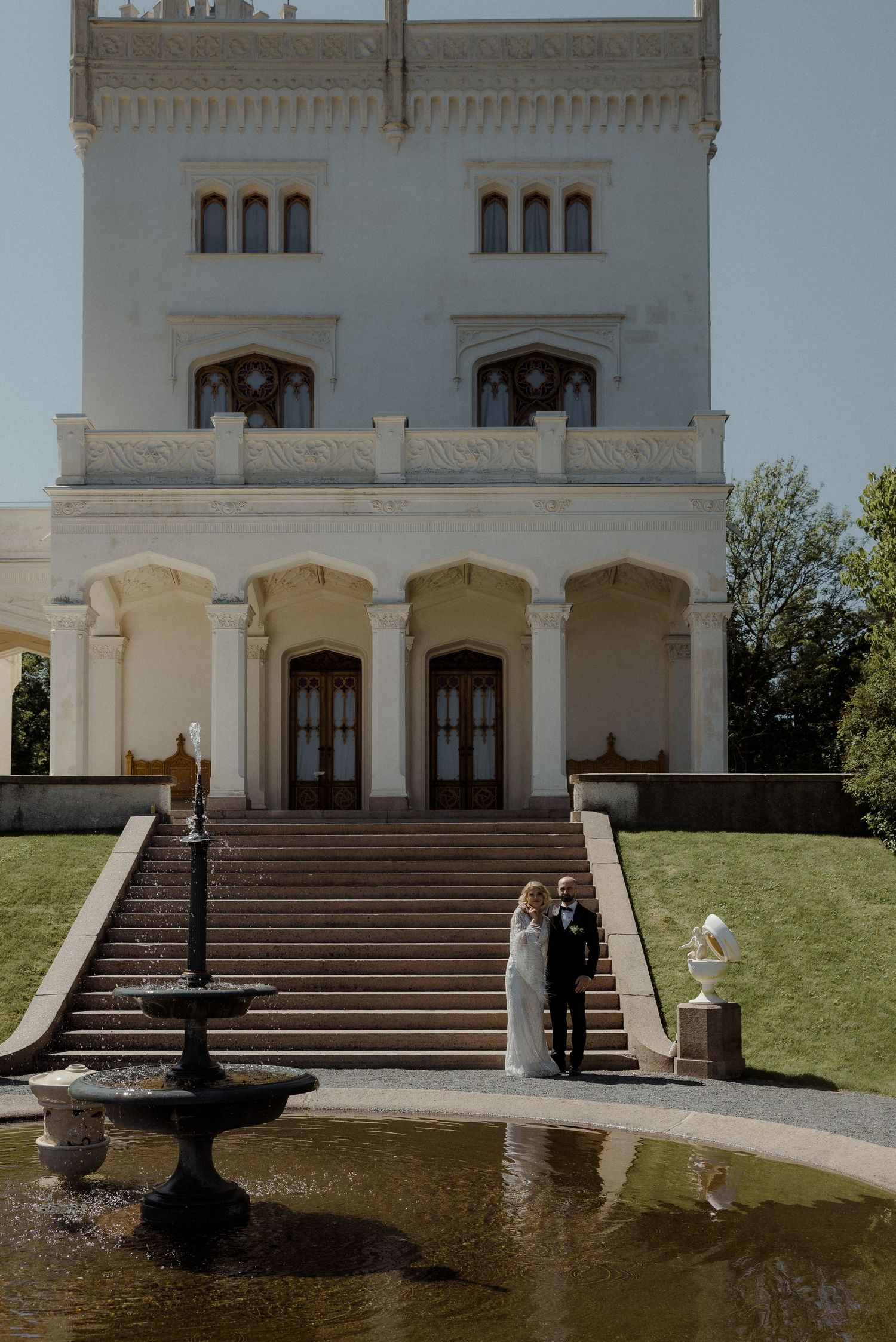 Bride and groom stand on grand staircase of elegant white castle venue with fountain in foreground.