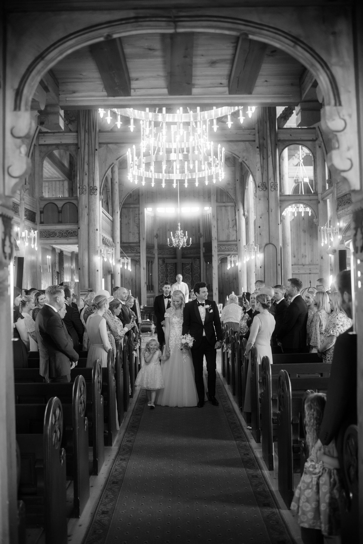 Black and white photo of a newlywed couple walking down the aisle of an elegant chandelier-lit church.