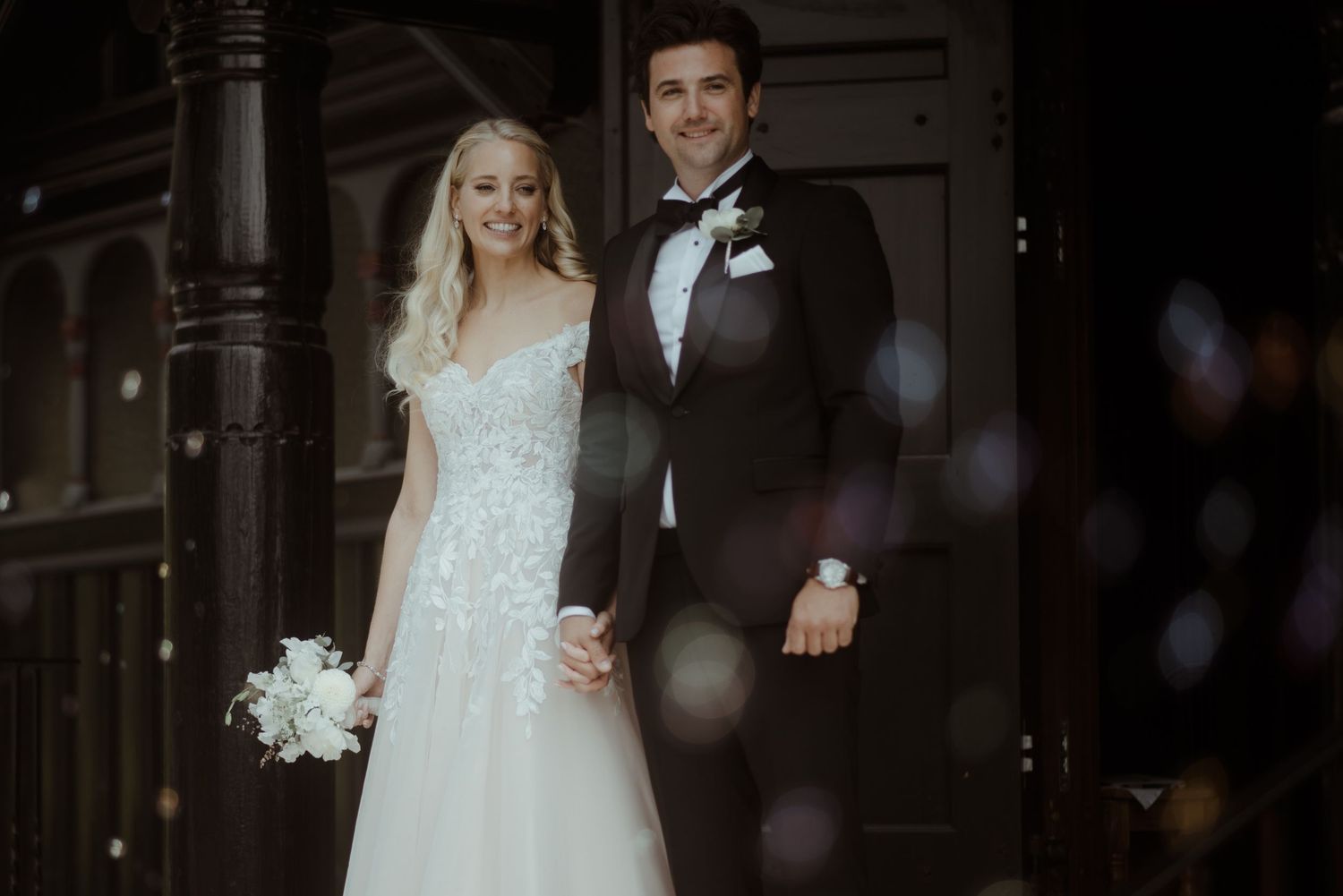 Bride and groom holding hands smiling in wedding attire outside a dark wooden door.