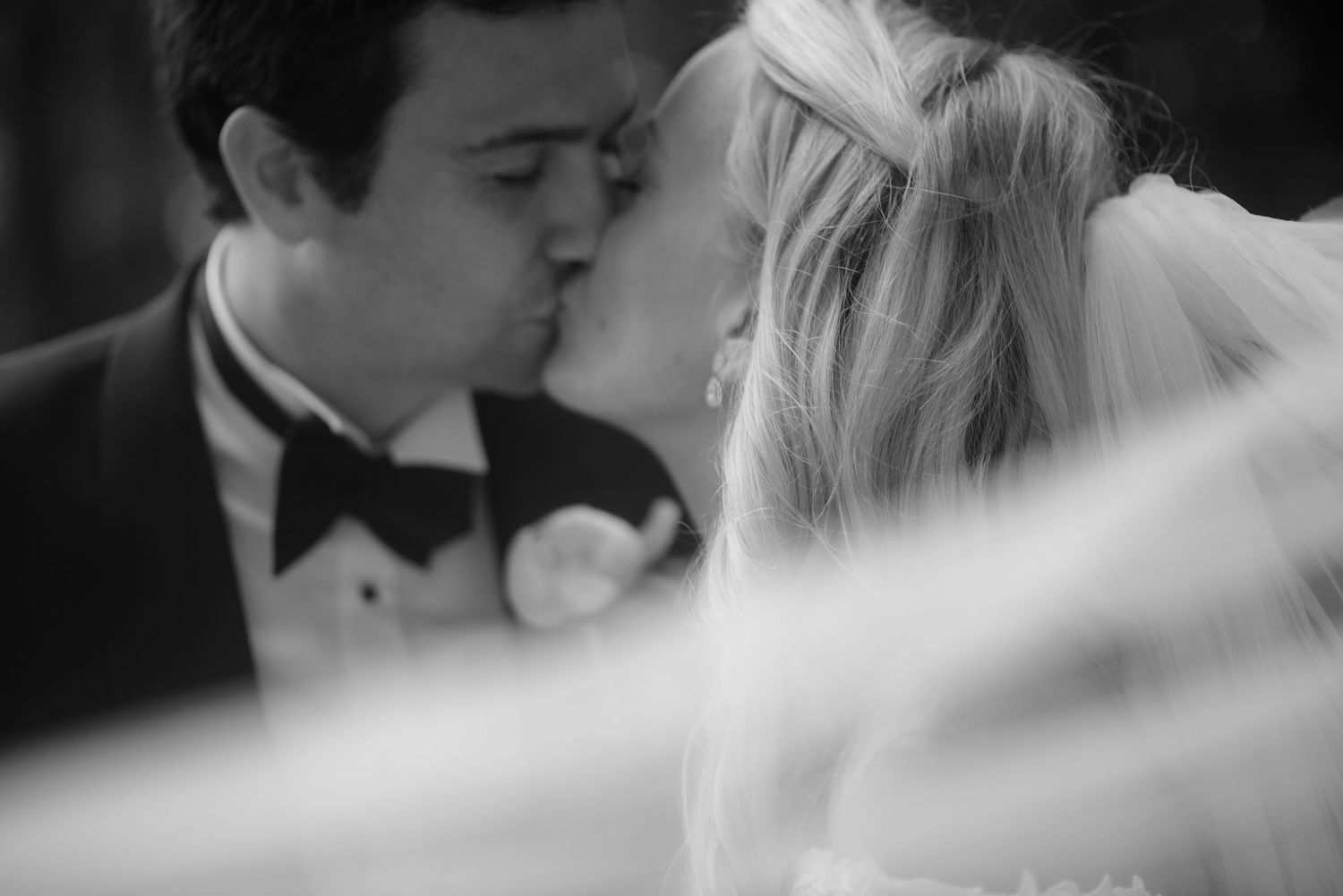 Black and white wedding photo of bride and groom sharing a romantic first kiss at their ceremony.