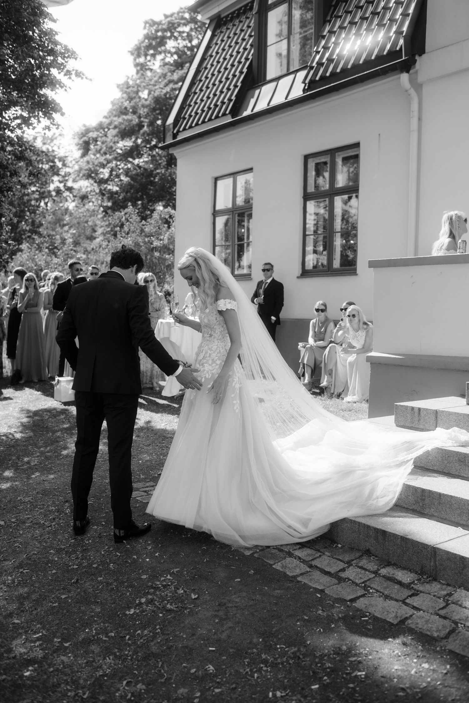 Black and white photo of bride and groom outside historic venue with flowing veil and wedding guests on steps.