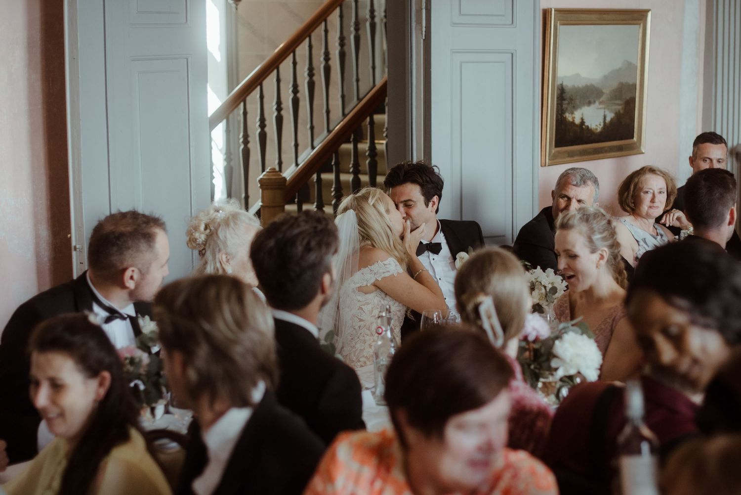Bride and groom share a kiss at elegant wedding reception with guests seated at decorated tables near a grand staircase.