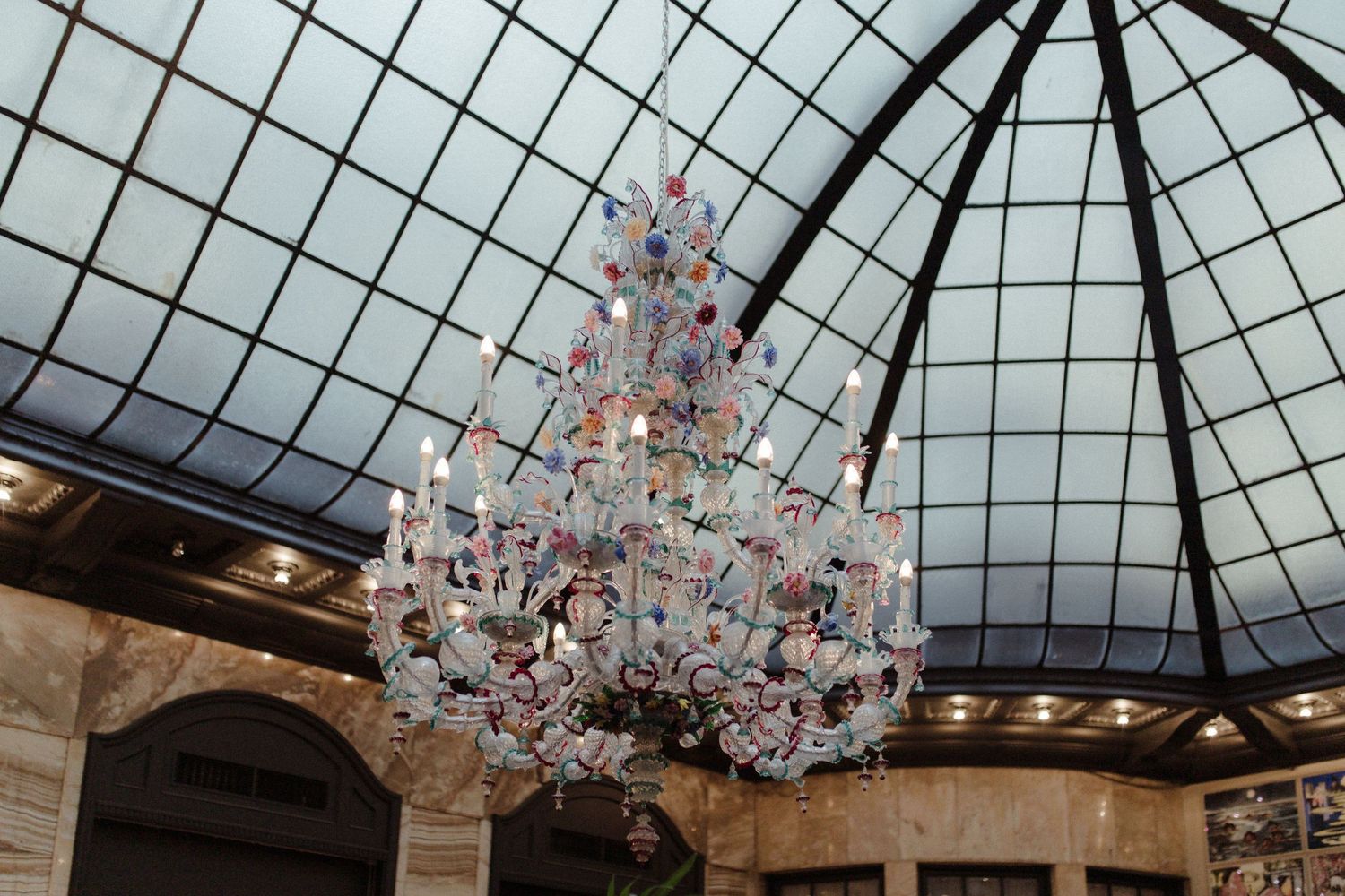 Ornate floral chandelier hanging beneath a domed glass ceiling in an elegant ballroom.