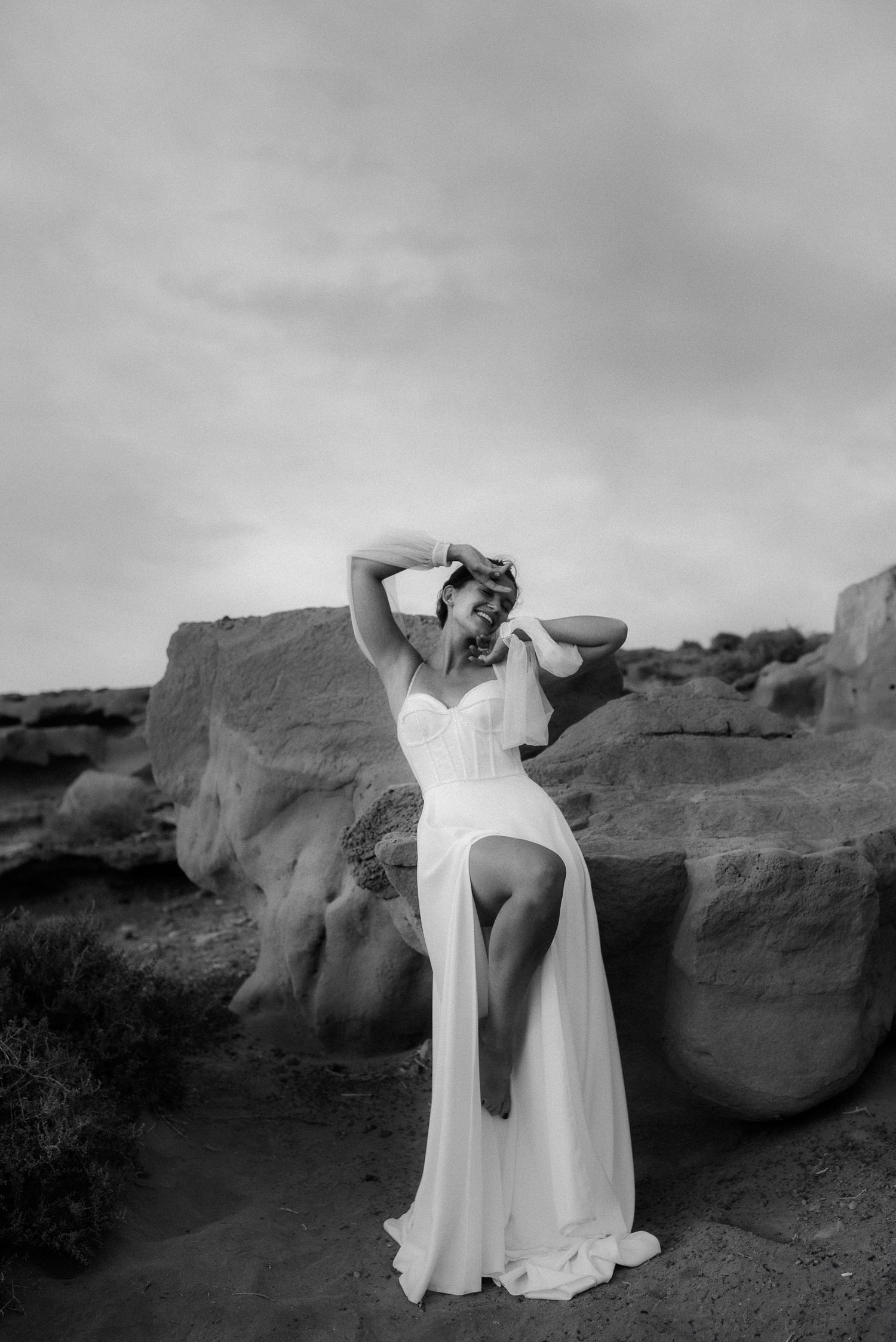 Black and white photo of a woman in a white corset gown posing against desert rocks under a cloudy sky.