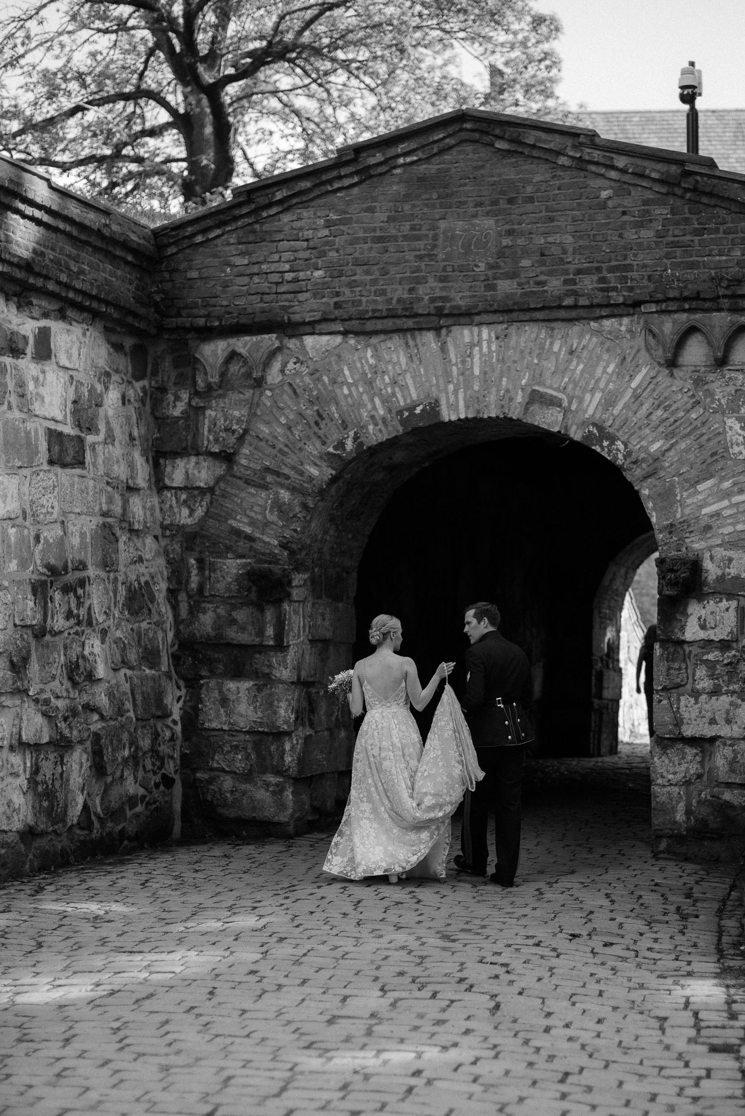 Black and white photo of a bride and groom holding hands walking through a stone archway on cobblestones.