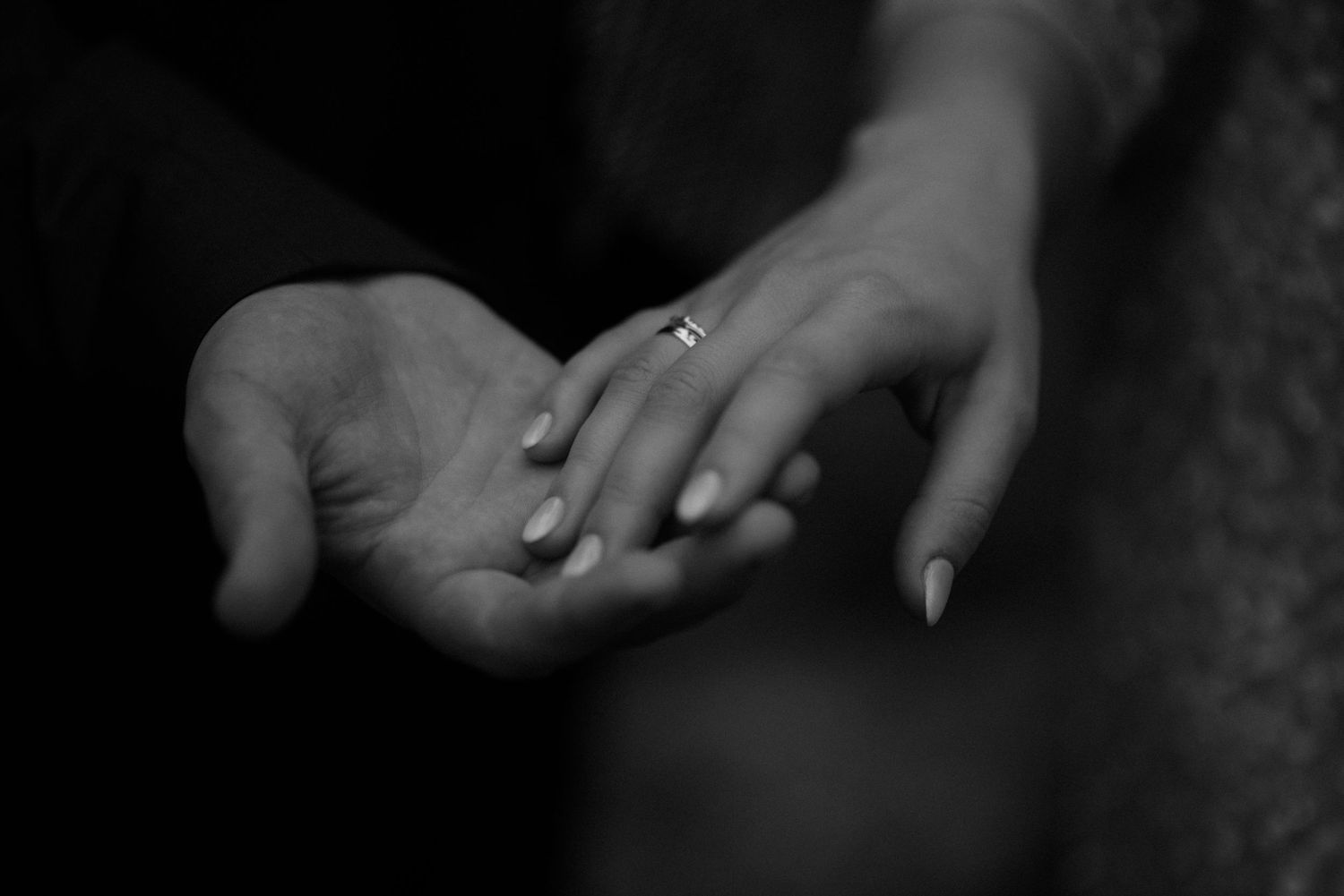 Black and white photo of two hands touching, one wearing a diamond engagement ring, symbolizing love and commitment.