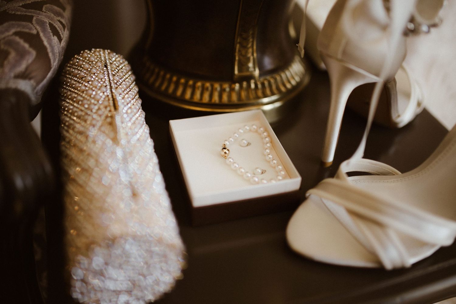 Elegant bridal accessories including pearl jewelry, white satin heels, and a beaded clutch arranged on a dark surface.