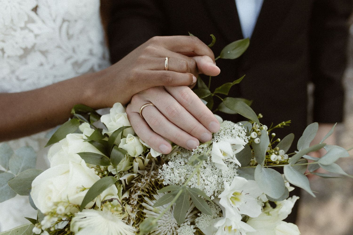 Newlywed couple holding hands over white floral wedding bouquet, both wearing gold wedding rings.