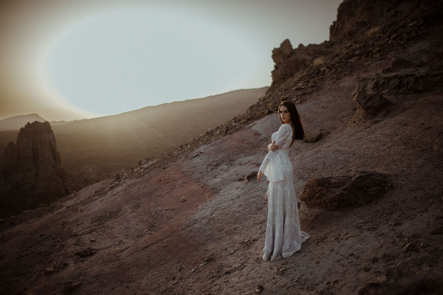 Pregnant woman in white lace gown poses on rocky desert hillside at golden sunset.