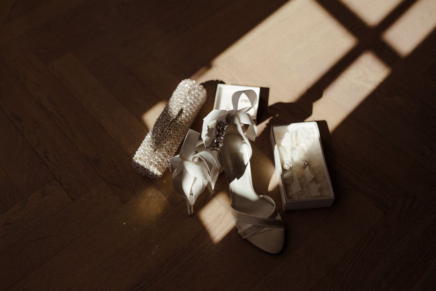 White wedding shoes and accessories arranged on a dark hardwood floor in warm sunlight.