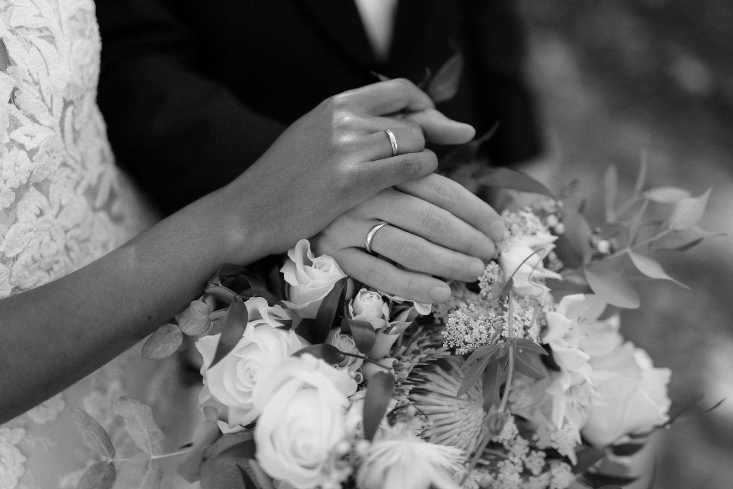 Black and white photo of bride and groom hands with wedding rings resting on a bridal bouquet.