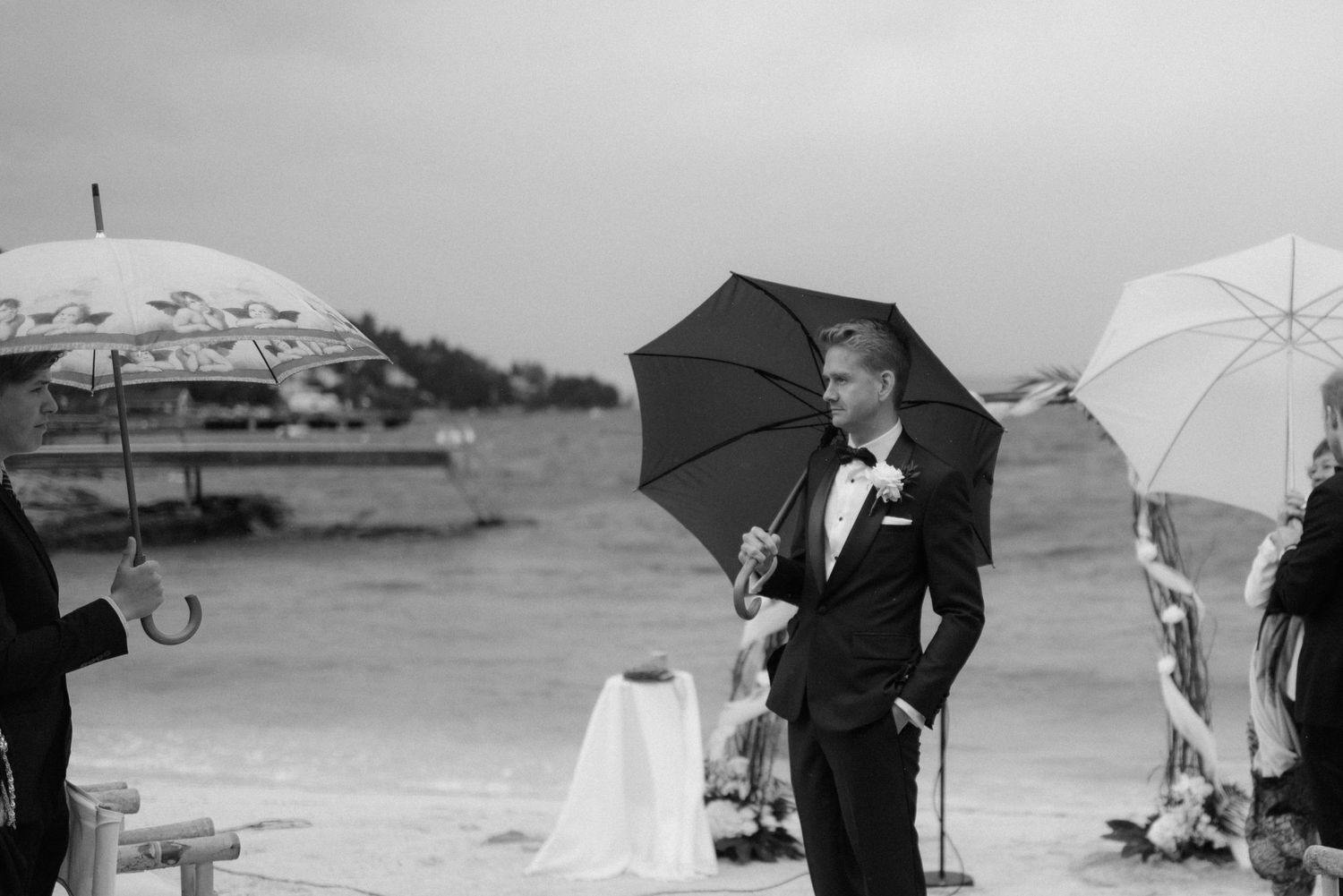 Groom in tuxedo holding umbrella at rainy beach wedding ceremony in black and white photo.