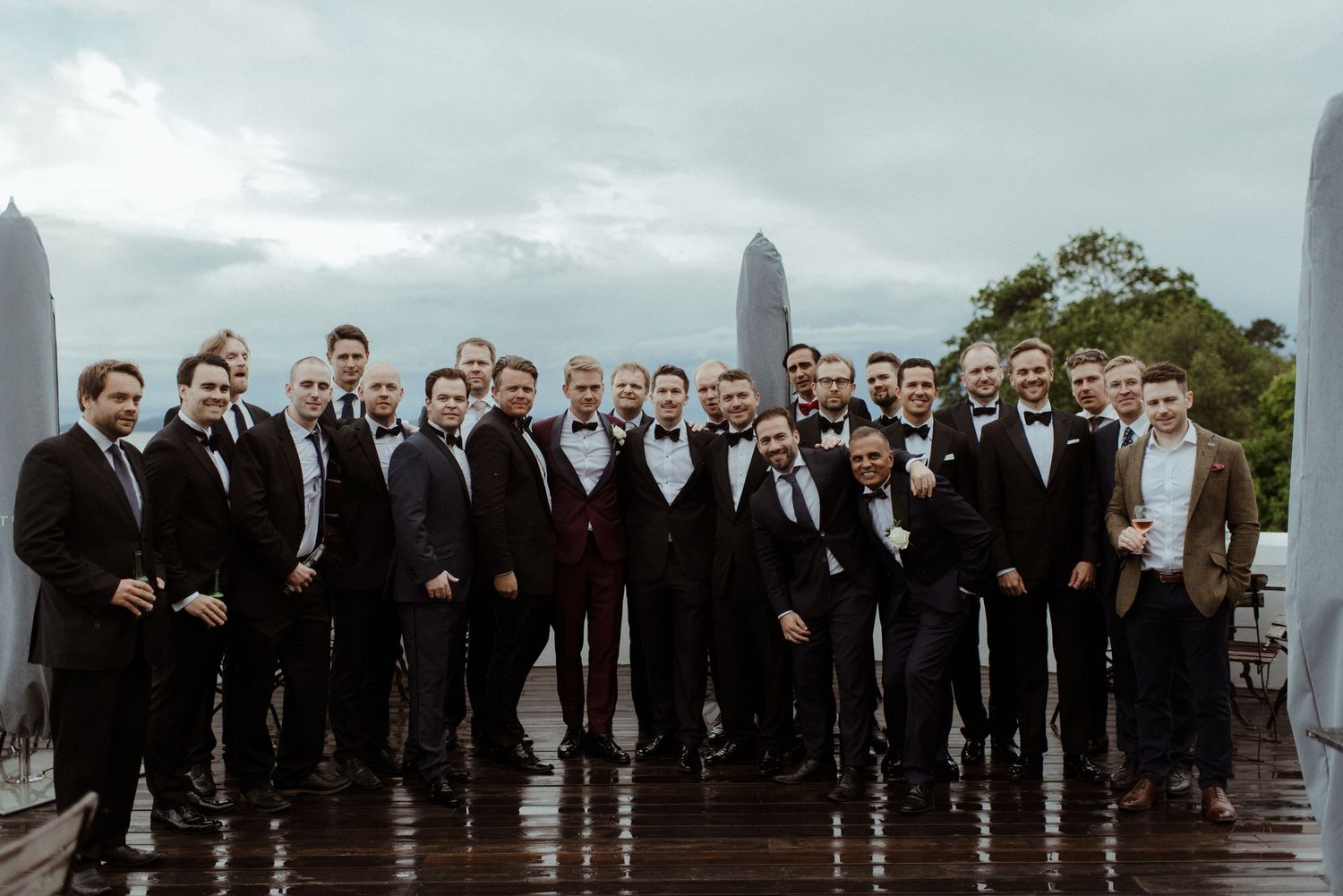 A large group of groomsmen in dark suits pose together on a rainy rooftop with a city skyline backdrop.
