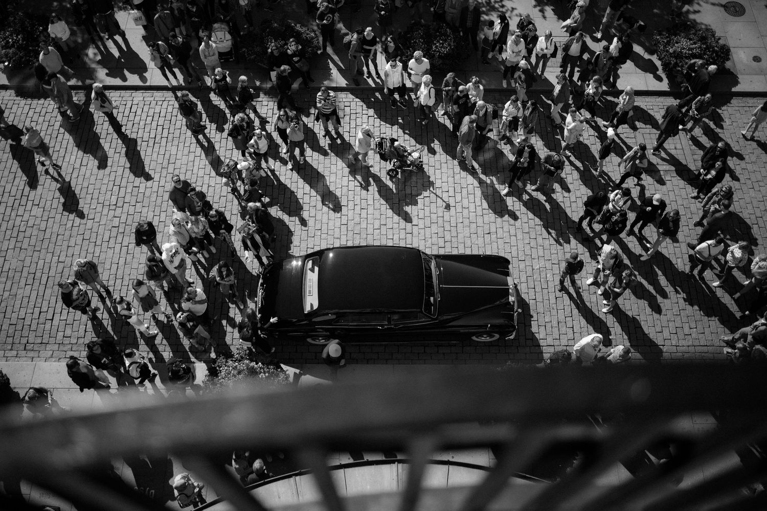 Aerial black and white photo of a dark car surrounded by crowds casting long shadows on pavement.