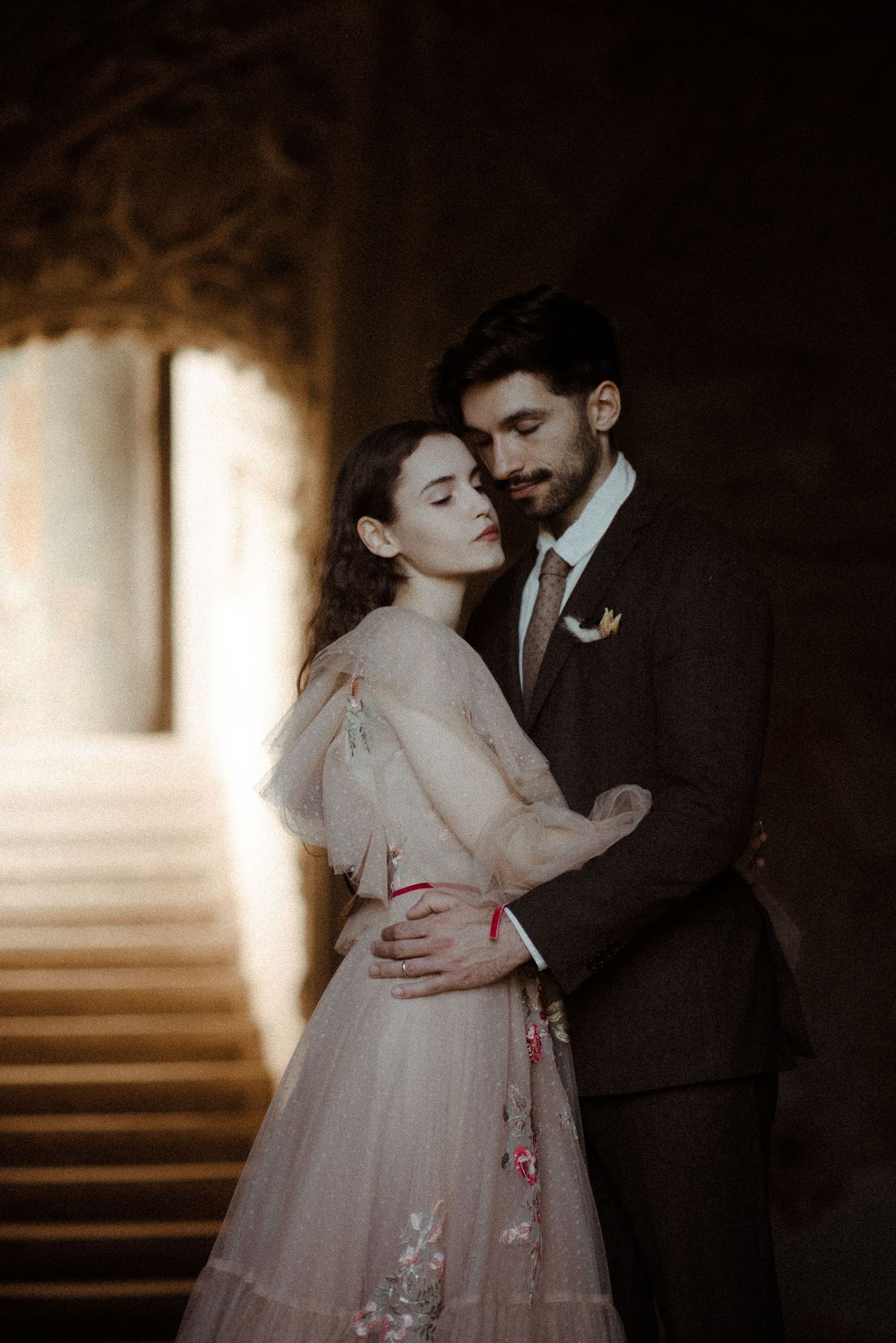 A couple in elegant wedding attire embrace tenderly in a warmly lit stone hallway, the bride in a blush floral gown.