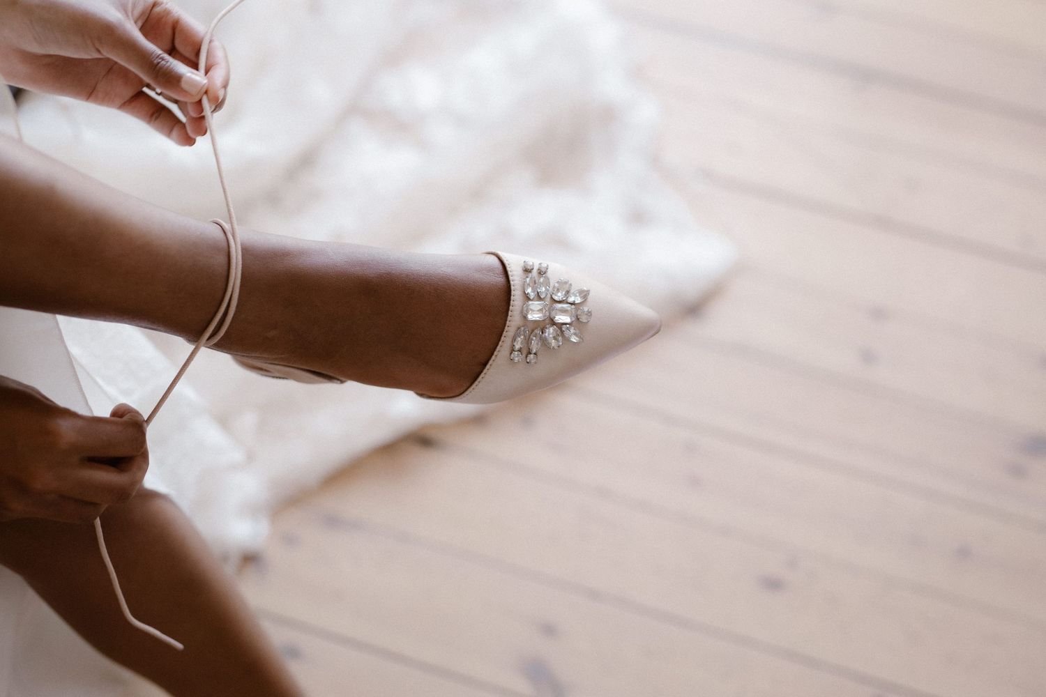 Bride tying ribbon on elegant white jeweled wedding heels on wooden floor.