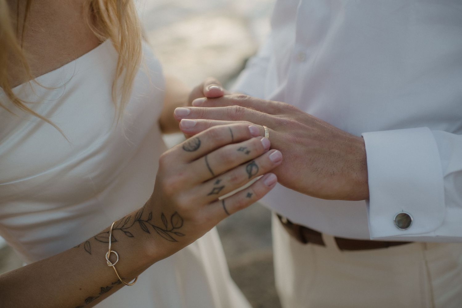 Bride and groom exchanging rings, bride wearing multiple rings and a delicate leaf tattoo bracelet on her wrist.