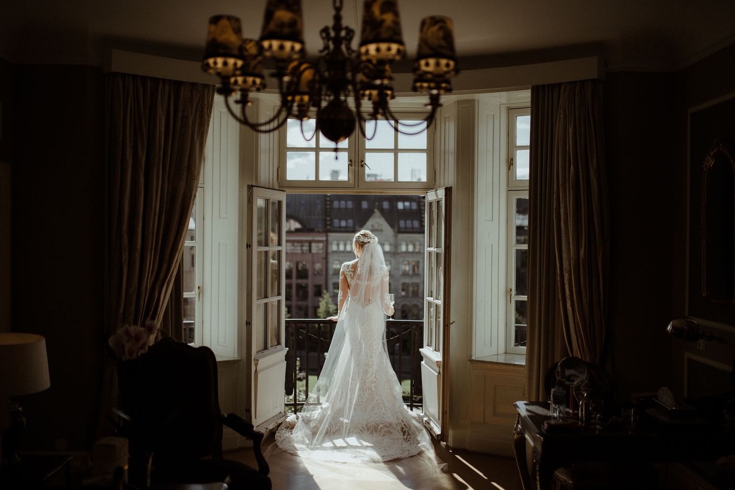 Bride in white gown and veil standing in open French doors of a luxury hotel room.