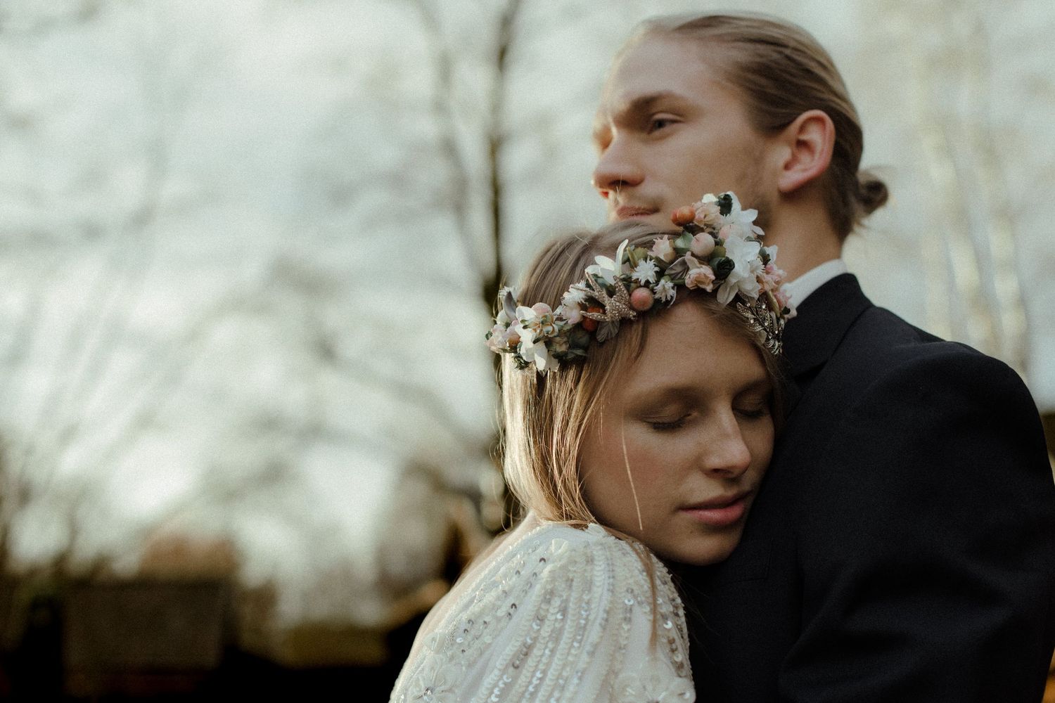 A bride wearing a floral crown leans against a groom in a black suit outdoors among bare trees.