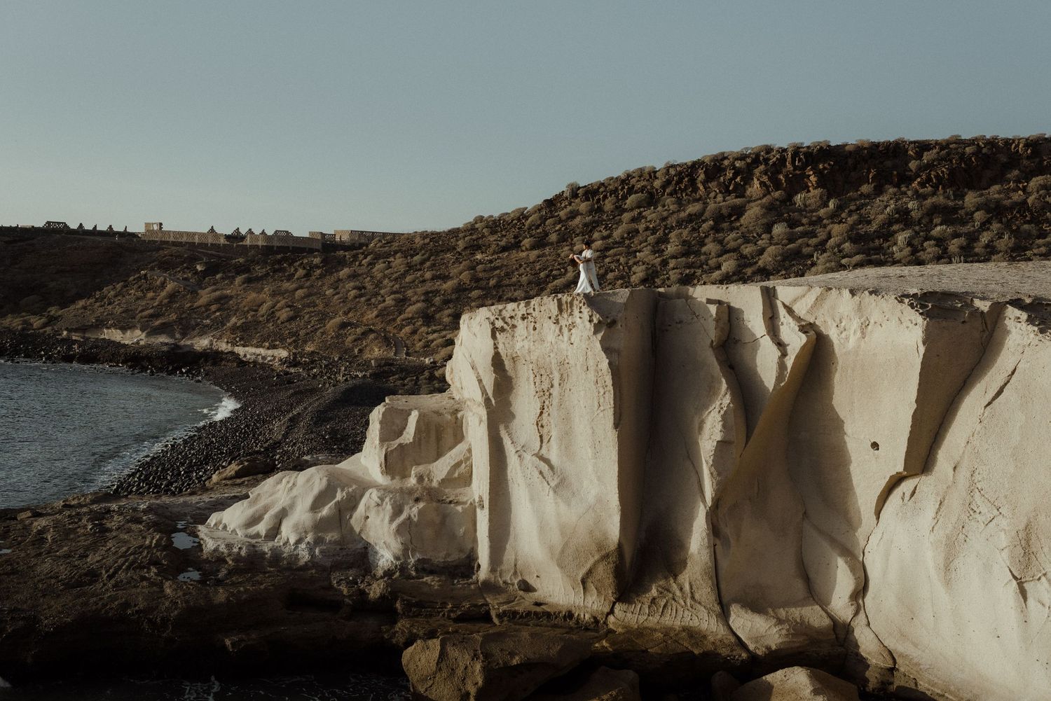 Person in white dress standing on dramatic white chalk cliffs overlooking the sea under a clear sky.