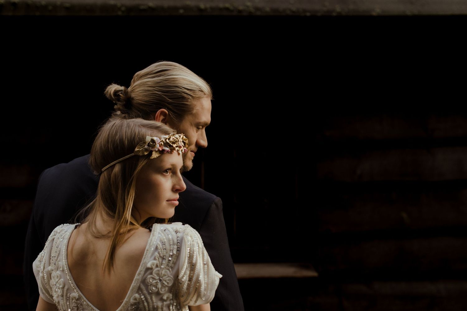 Bride in lace backless gown with floral crown leans against groom in dark suit in moody rustic barn setting.