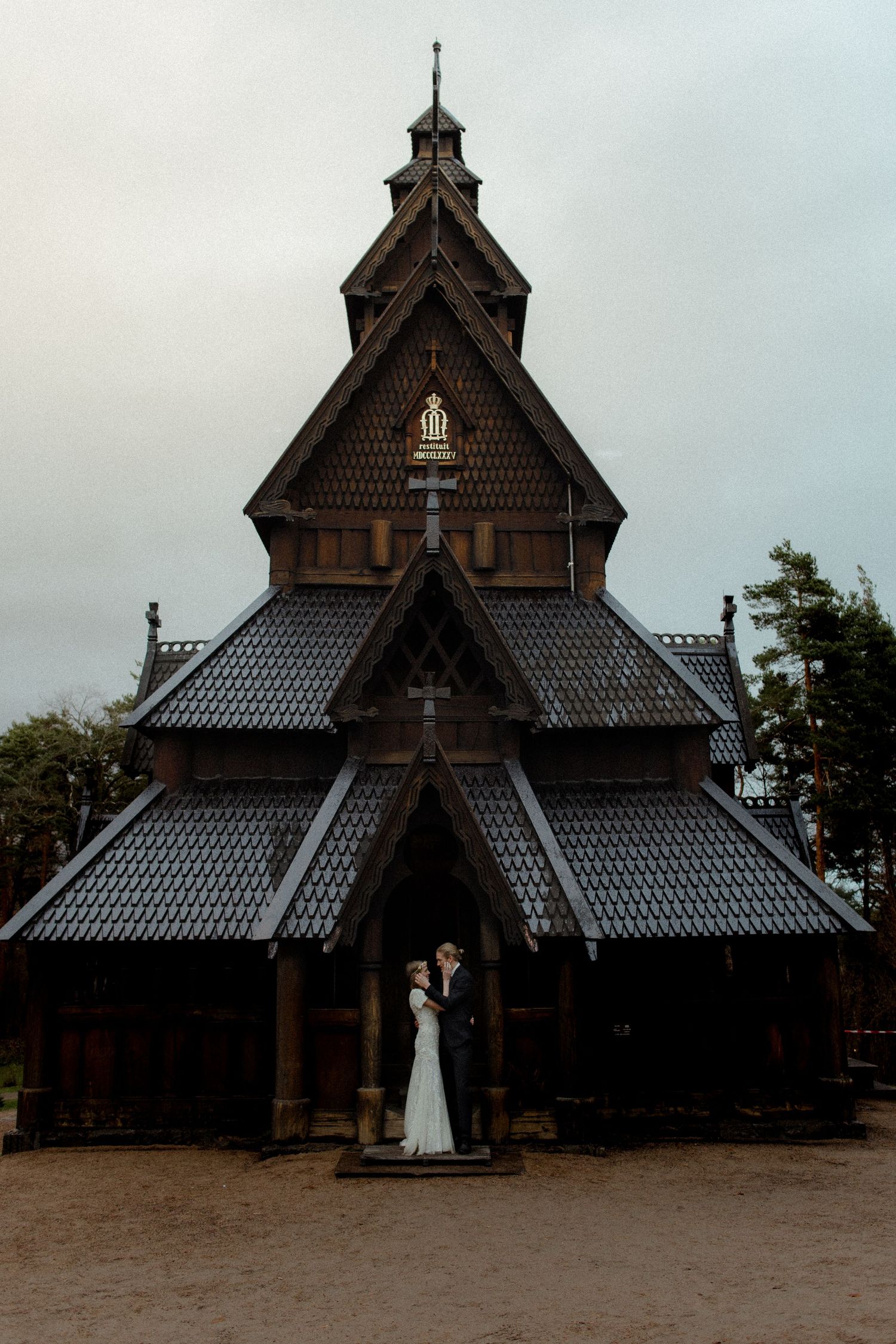 Wedding couple kissing before a dark wooden Norwegian stave church with ornate peaked roofs and cross details.