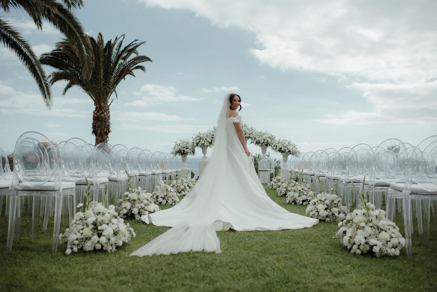 Bride in elegant white gown with long train stands at outdoor wedding ceremony aisle with white floral arrangements and ghost chairs.