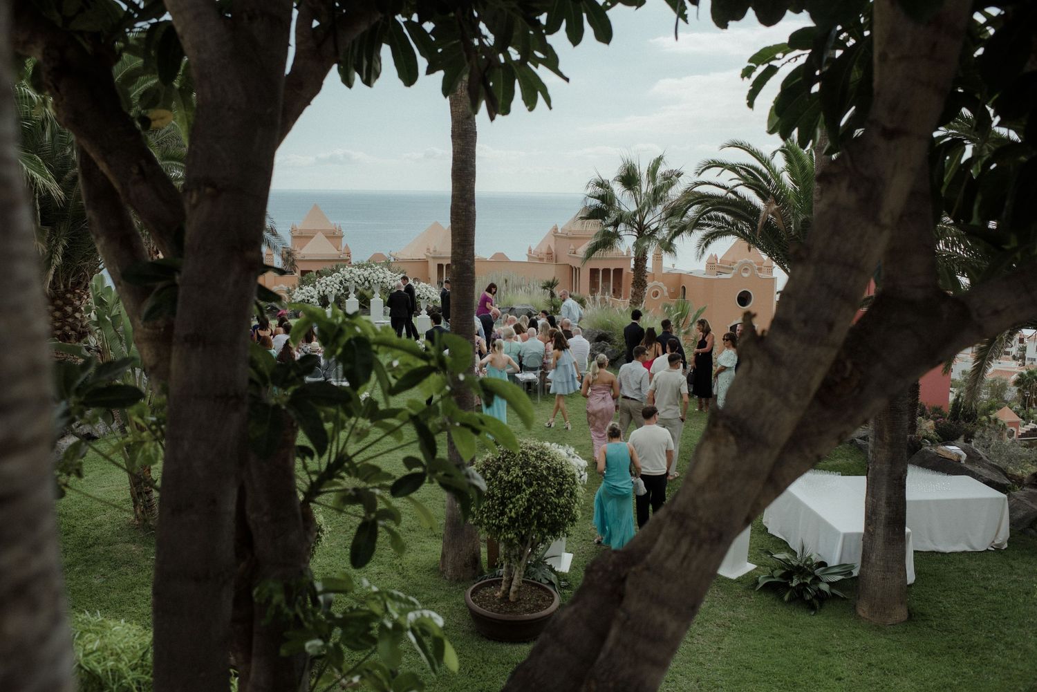 Outdoor wedding reception guests gather in lush garden with ocean view and palm trees.