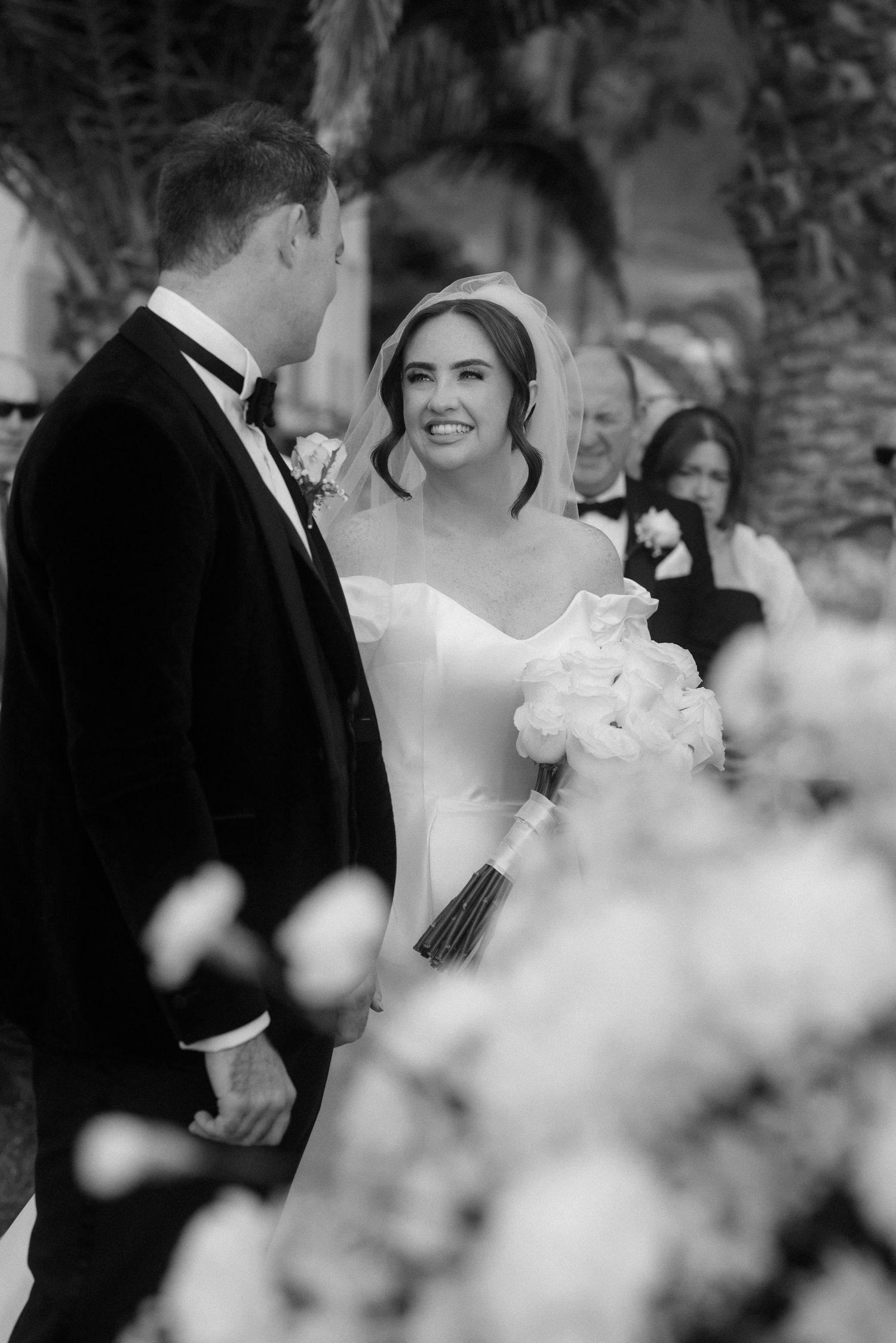 Black and white wedding photo of smiling bride in veil holding bouquet facing groom in tuxedo at outdoor ceremony.