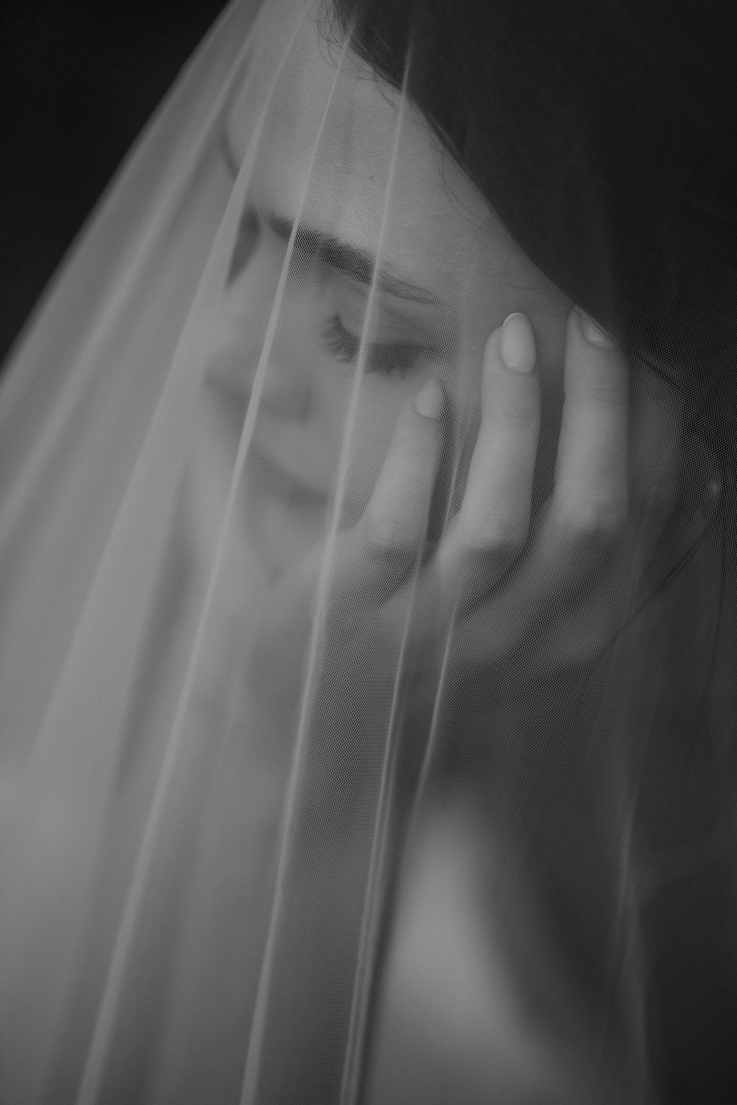 Black and white portrait of a woman behind a sheer veil, hand raised, eyes closed, in a dreamy artistic composition.
