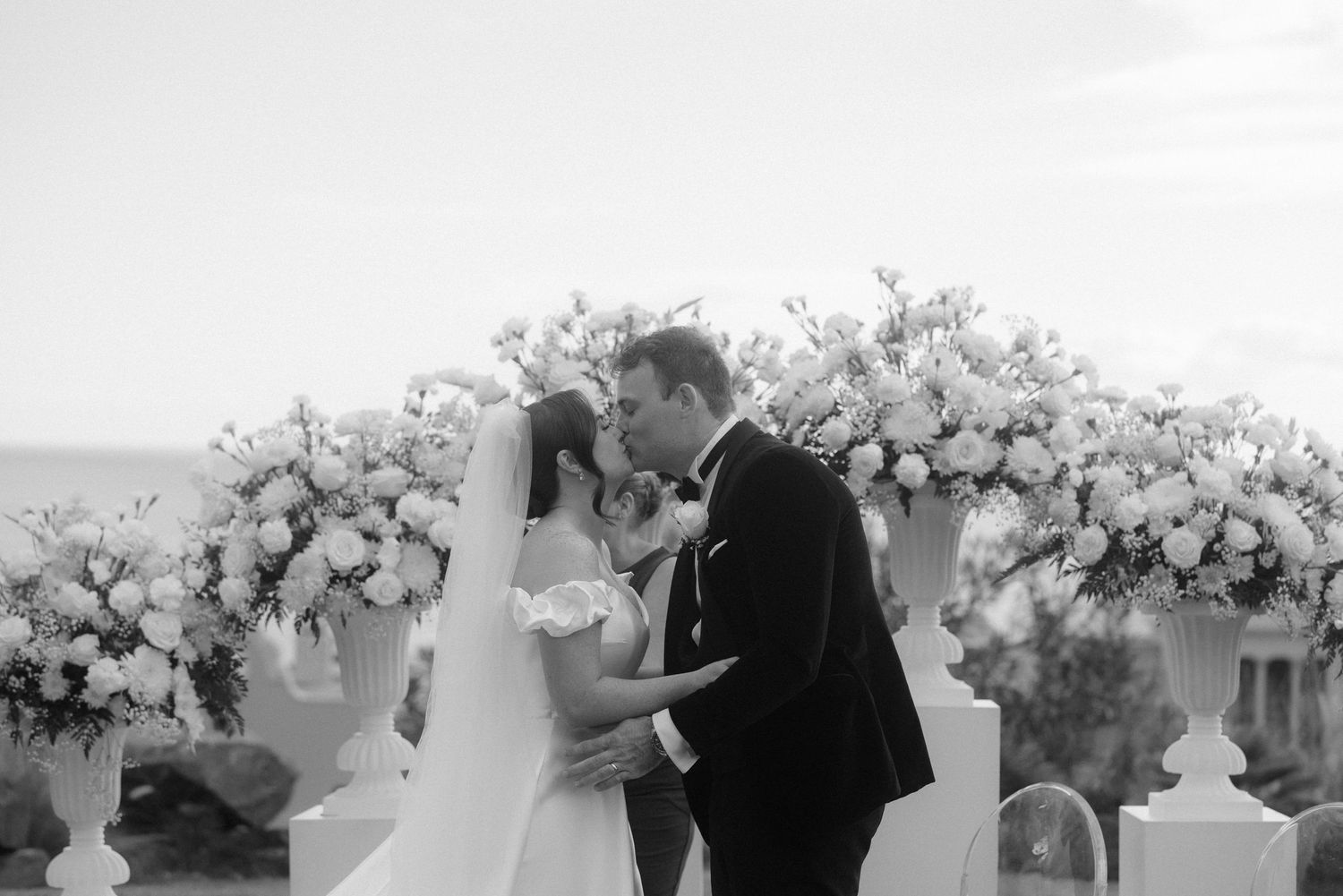 Black and white wedding photo of bride and groom kissing at outdoor ceremony with floral arrangements.