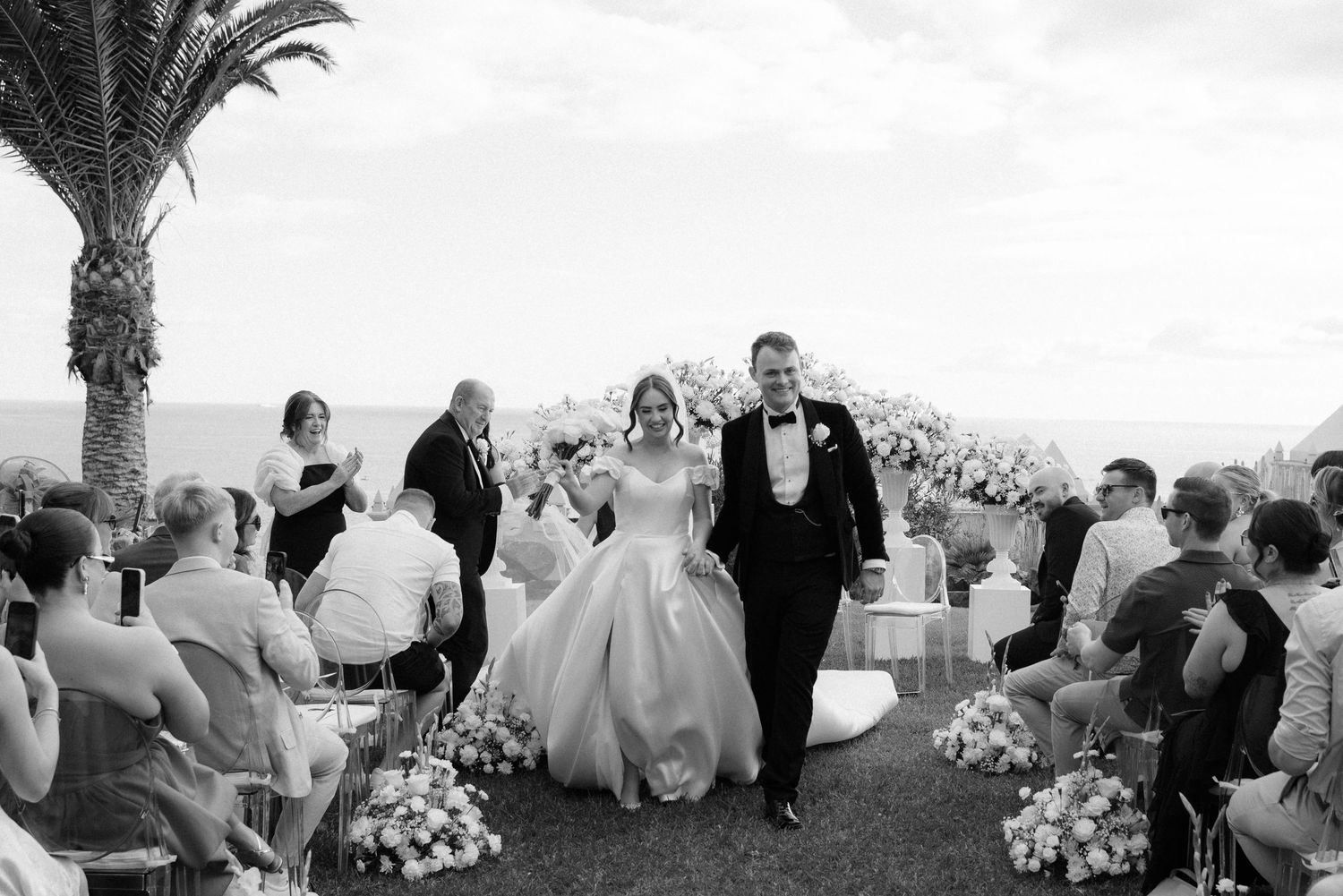 Bride and groom walk down the aisle at an outdoor wedding ceremony with ocean views and floral arrangements.