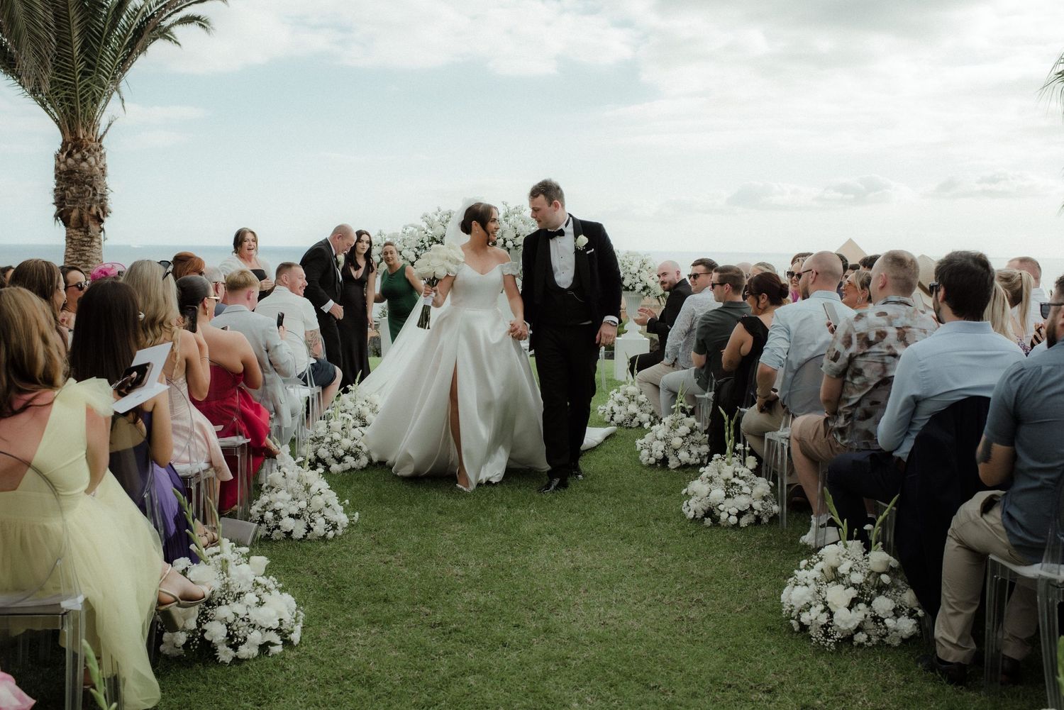 Bride and groom walk down outdoor aisle lined with white floral arrangements at elegant seaside wedding ceremony.
