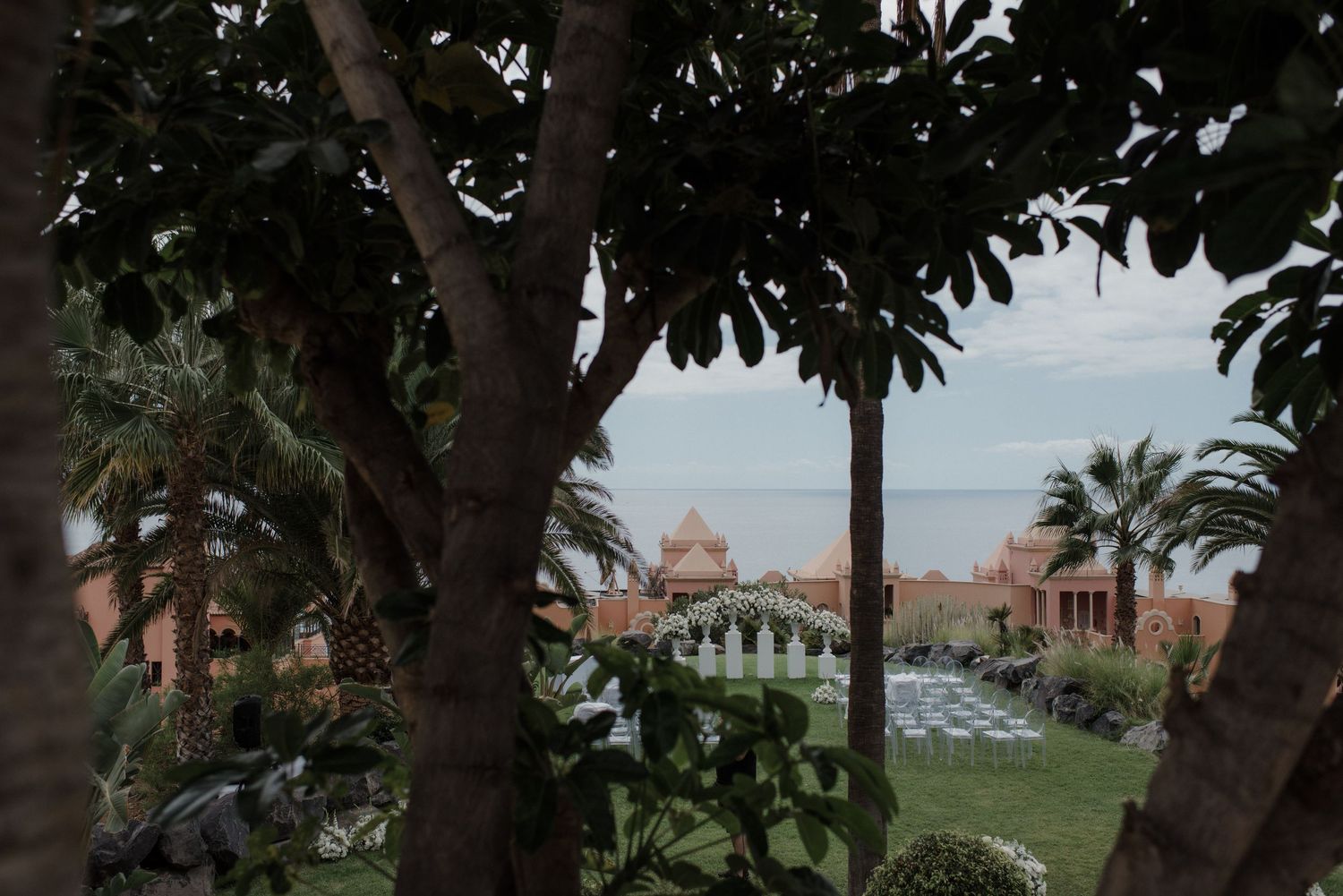Lush tropical garden with palm trees framing a luxury resort and ocean view under an overcast sky.