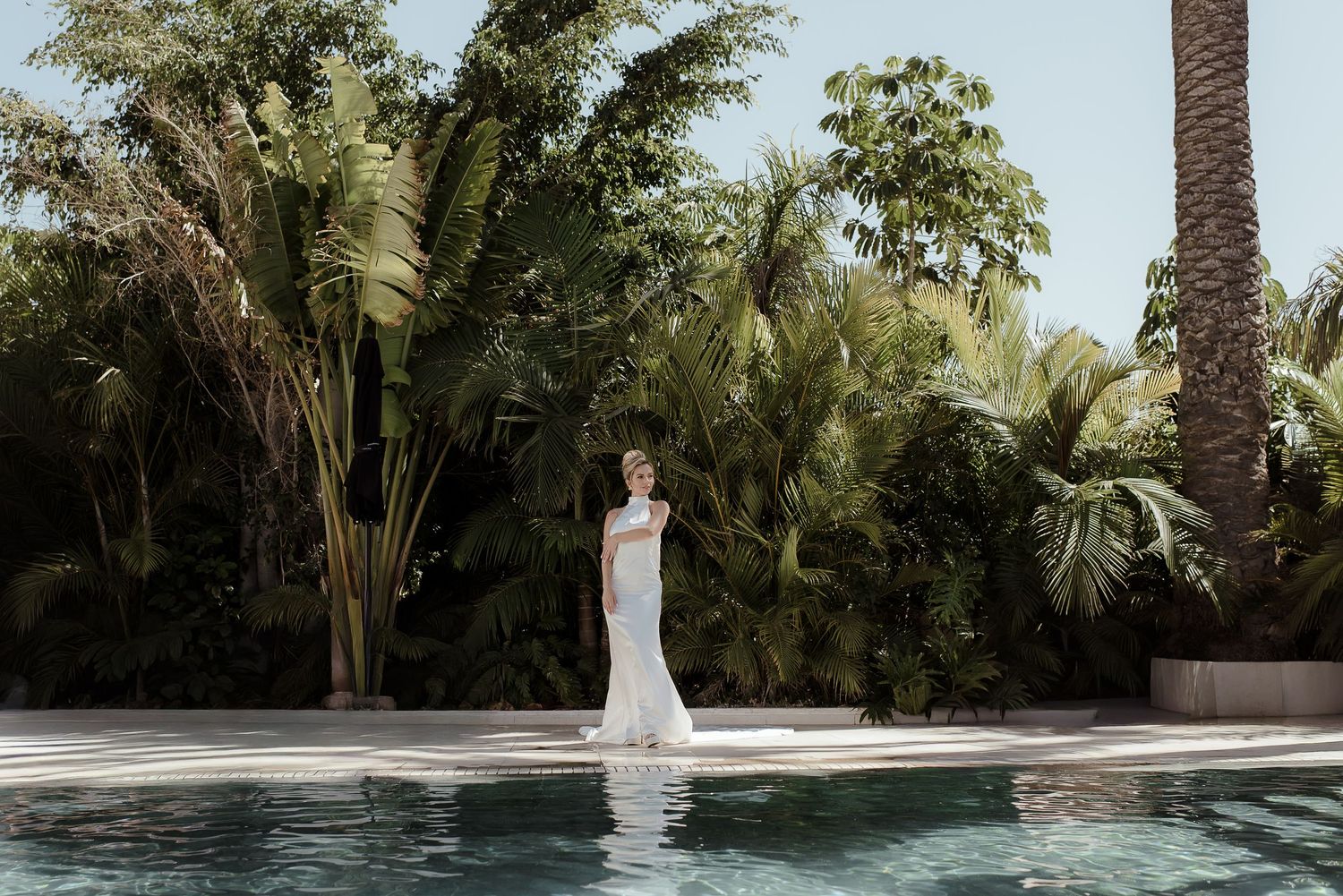 Bride in white wedding gown standing poolside surrounded by lush tropical palm trees and greenery.