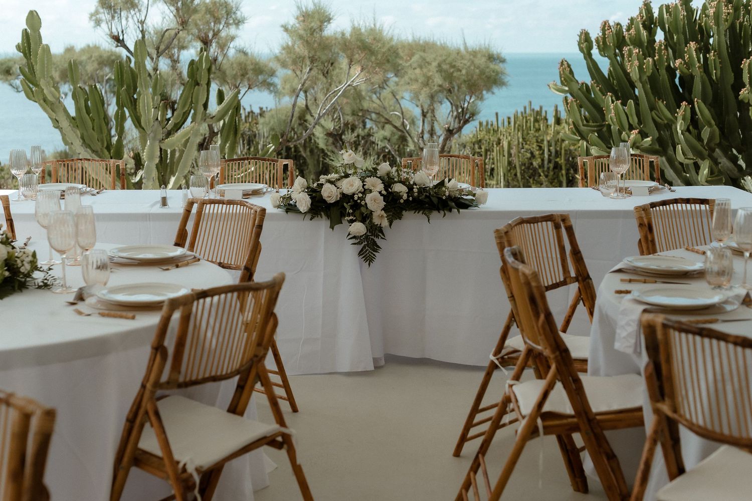 Elegant outdoor wedding reception with wooden chairs, white tables, floral centerpiece, and cacti overlooking the sea.