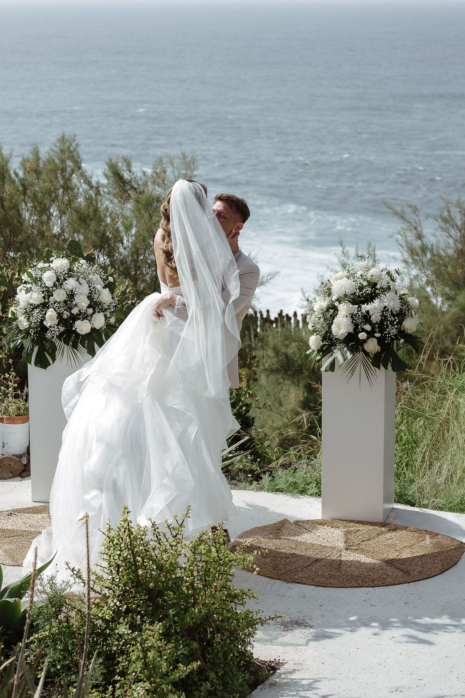 Bride and groom share a kiss at an outdoor ocean-view wedding ceremony with white floral arrangements.