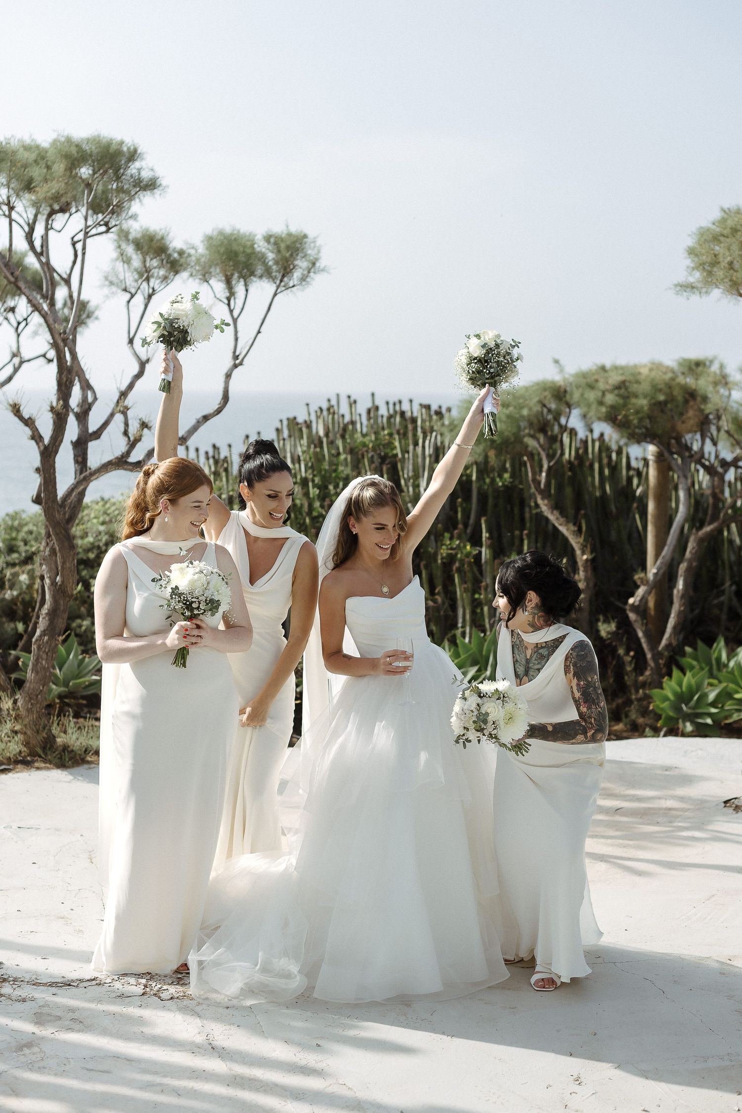 Bride and bridesmaids in white gowns celebrate outdoors, raising bouquets of white flowers with ocean and trees behind.