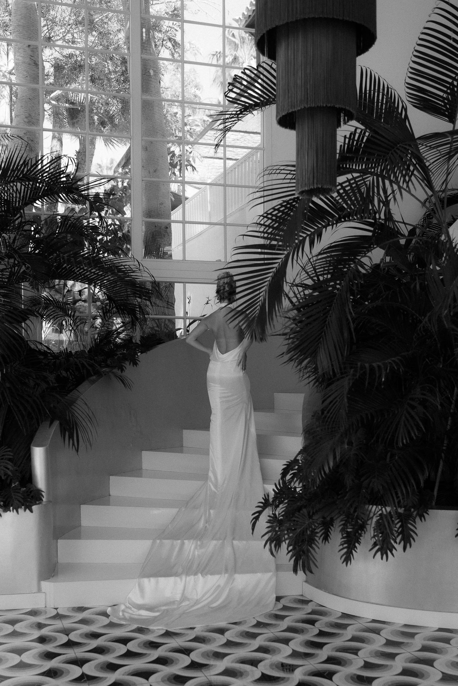 Elegant bride in a fitted white gown with train stands on stairs amid tropical palms in black and white photo.